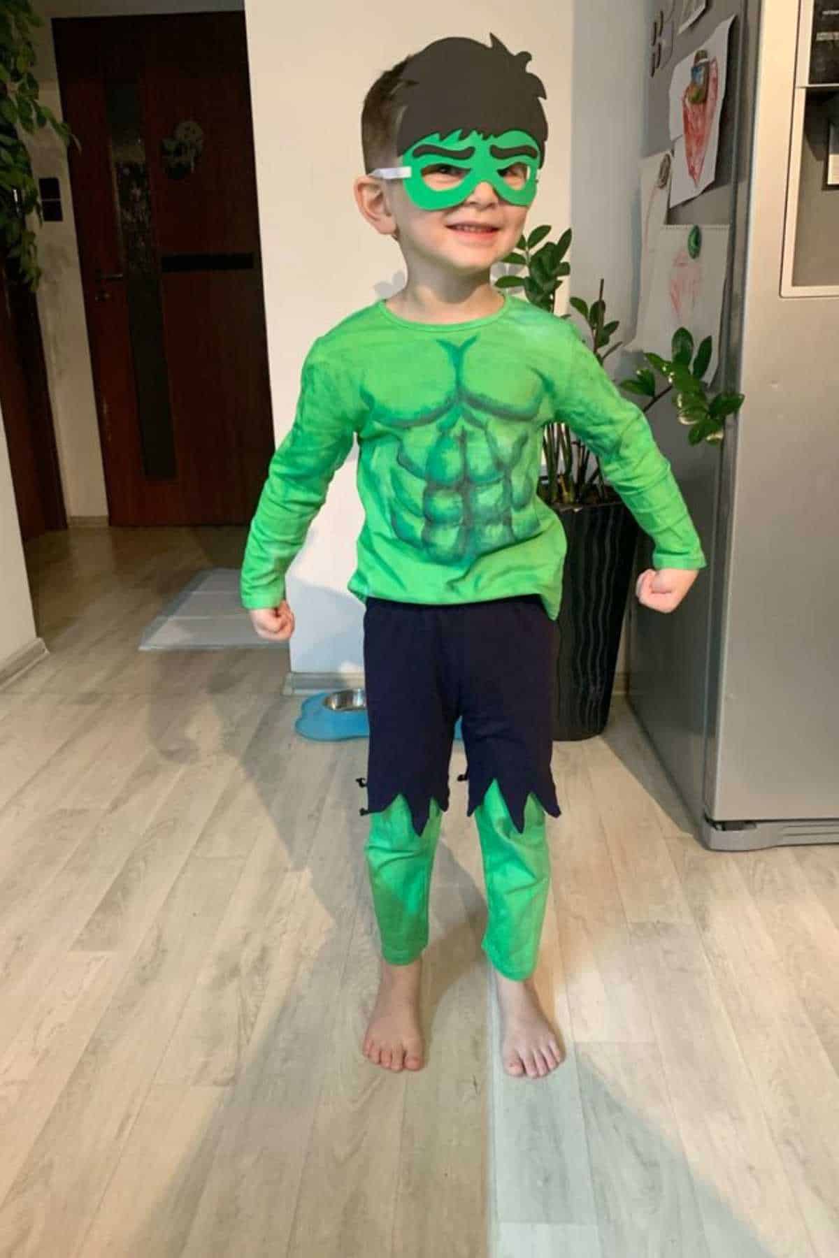 Young child wearing a green Hulk costume with a matching mask, standing barefoot on a wooden floor in a home setting, smiling and posing with fists clenched.