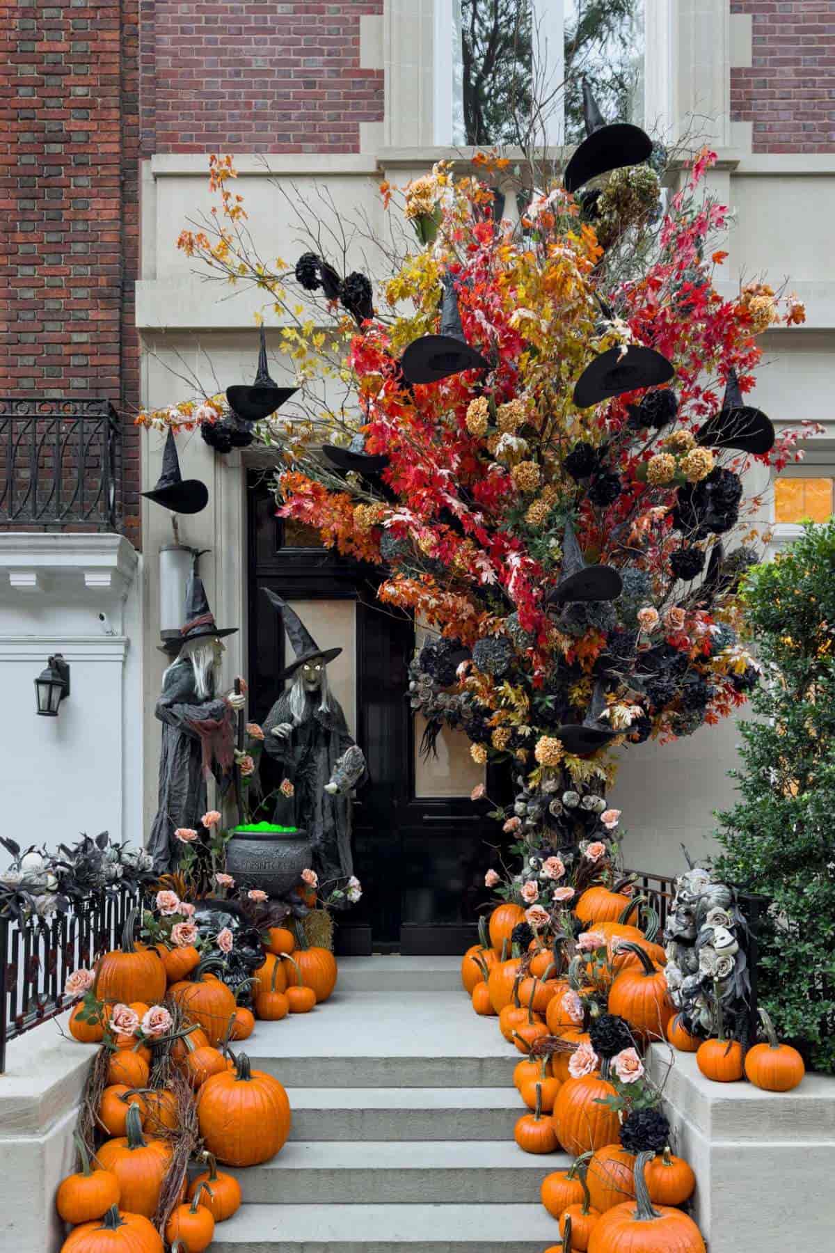 Front steps decorated for Halloween with pumpkins, flowers, witch statues, and black witch hats hanging from a colorful tree in front of a brick building.