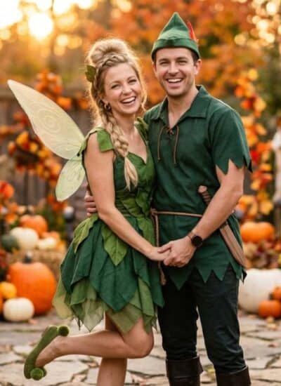 Two adults dressed as fairy tale characters stand smiling together in a fall-themed garden decorated with pumpkins, flowers, and autumn leaves.
