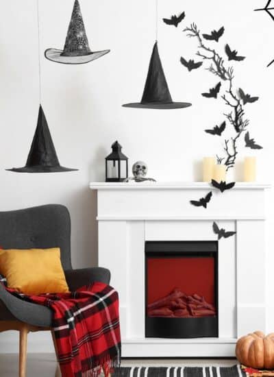 A living room decorated for Halloween with pumpkins, a faux fireplace, skulls, witch hats, bats, spider webs, ghosts, and a cauldron, plus a gray chair with a red plaid blanket and yellow pillow.