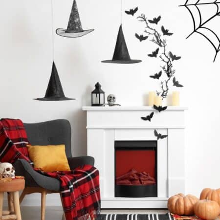 A living room decorated for Halloween with pumpkins, a faux fireplace, skulls, witch hats, bats, spider webs, ghosts, and a cauldron, plus a gray chair with a red plaid blanket and yellow pillow.