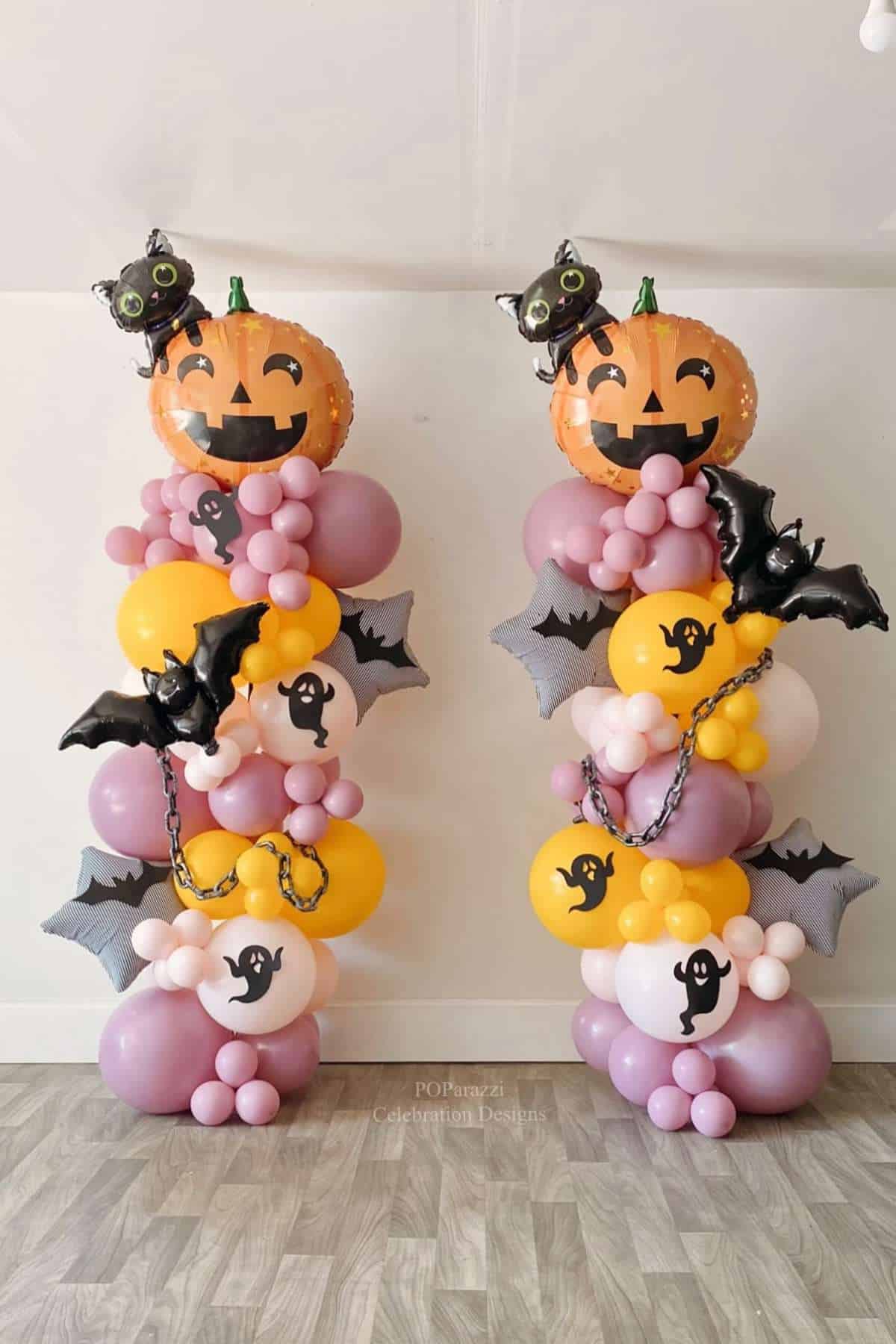 Two balloon towers with pumpkin, bat, and ghost designs stand on a wood floor, decorated for Halloween with pink, yellow, and black colors.