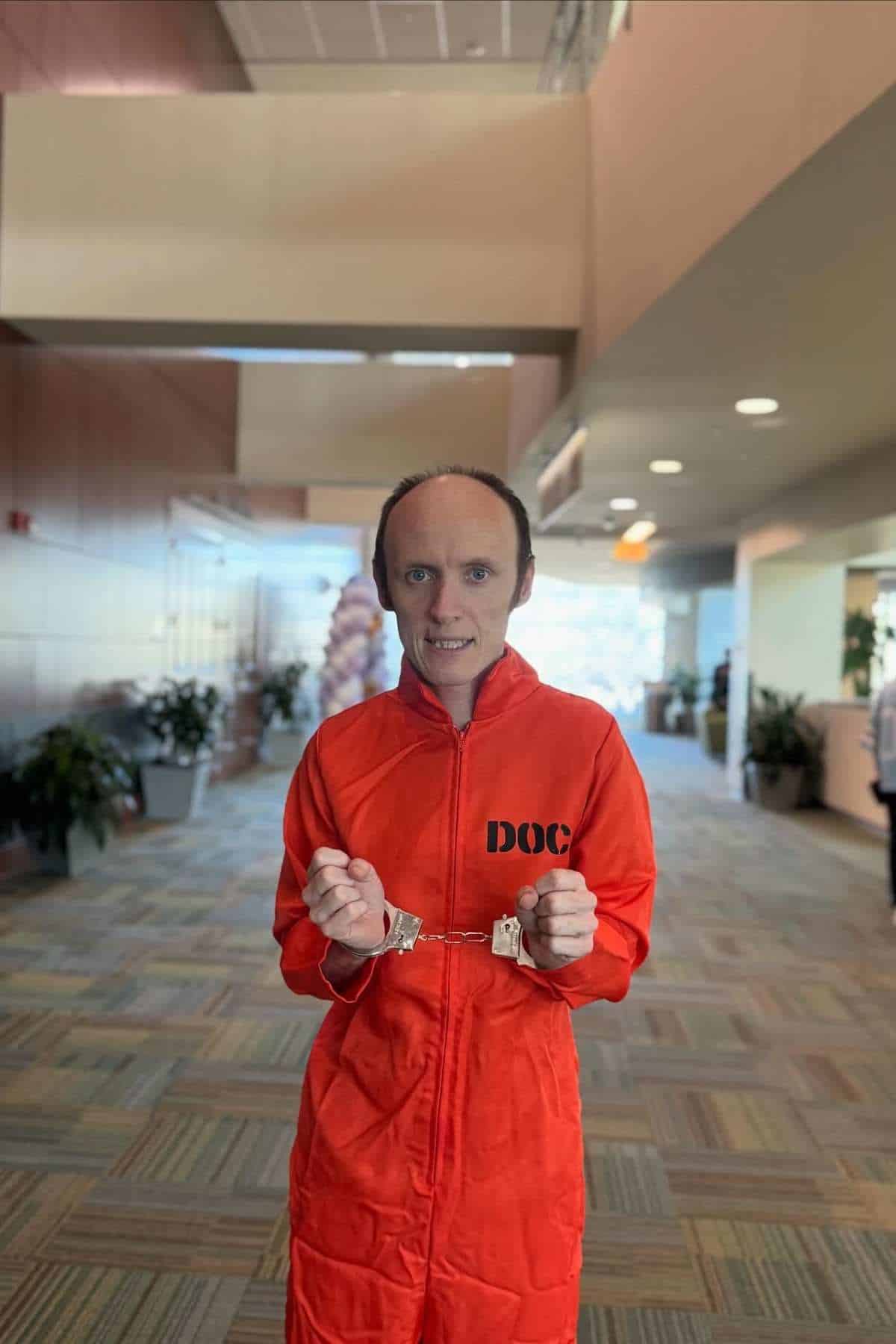 A person in an orange jumpsuit marked "DOC" stands indoors, wearing handcuffs and looking at the camera.
