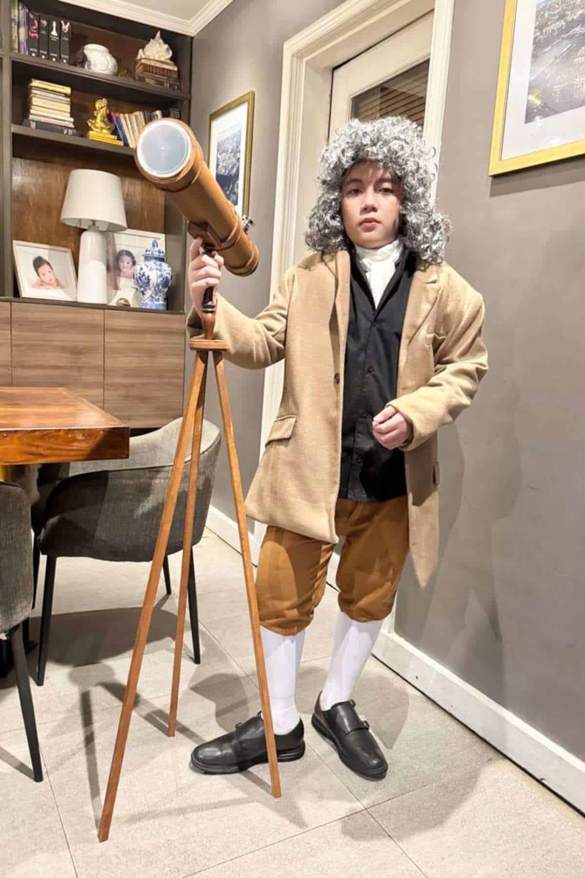 A child dressed in historical clothing and a gray wig stands indoors next to a wooden telescope on a tripod.