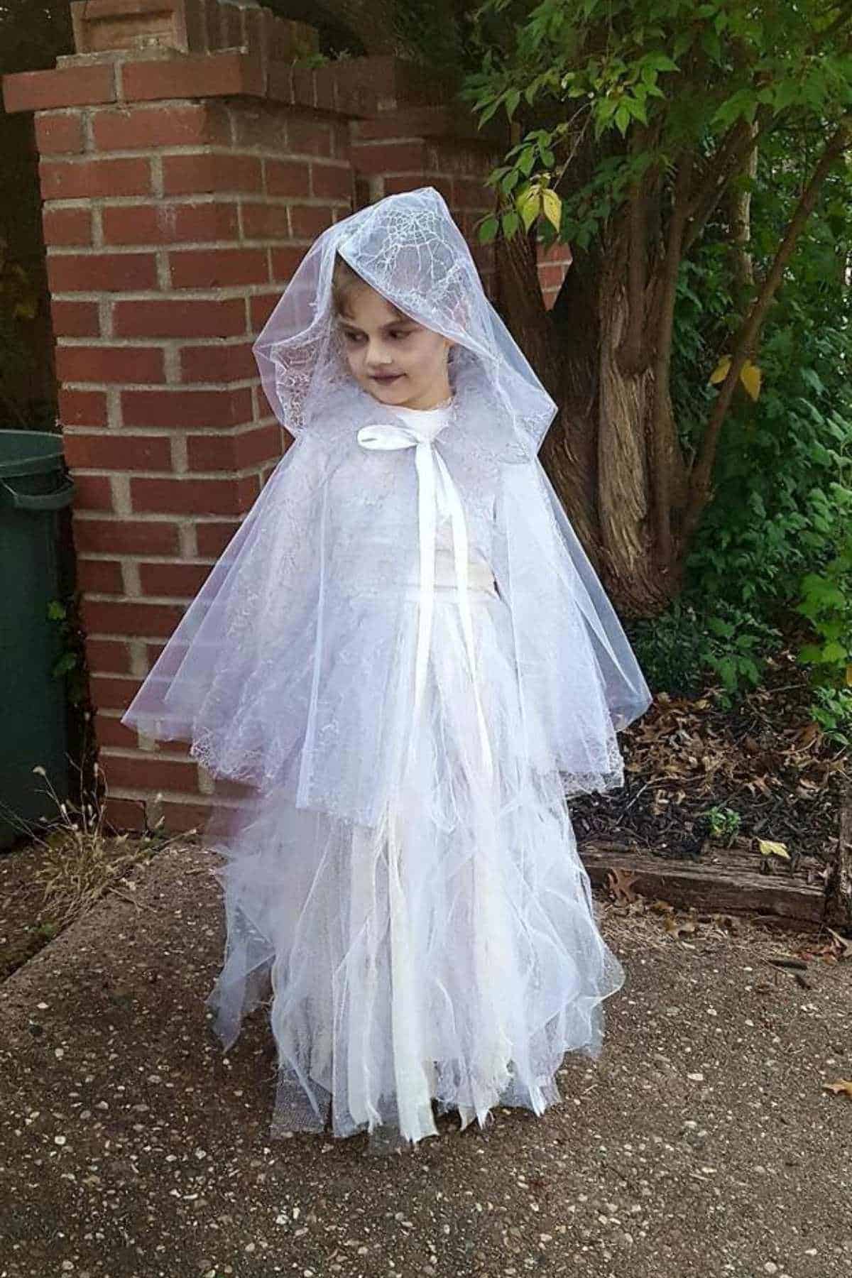 A young child stands outside wearing a white lace dress and a matching hooded cape, resembling a ghost or bride.