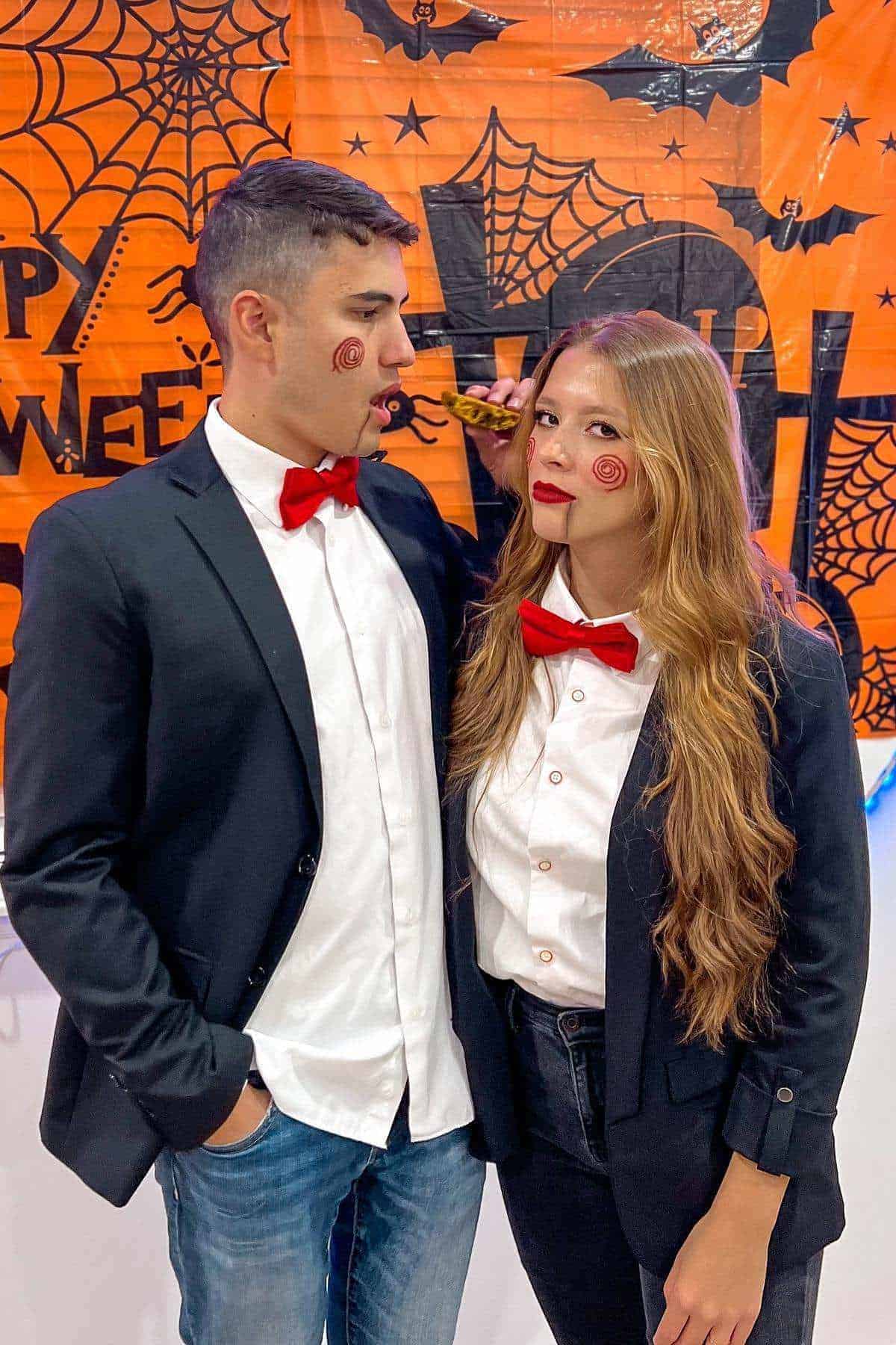 Two people dressed in matching black suits, white shirts, and red bow ties with painted spiral cheek makeup, standing in front of a Halloween-themed backdrop.
