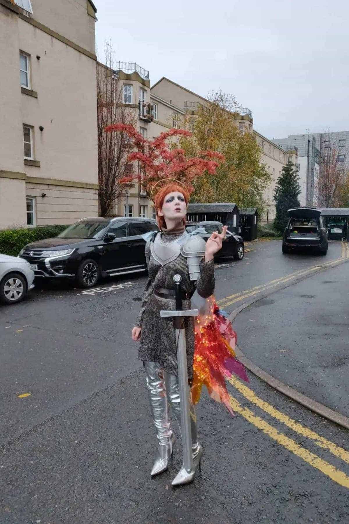 Person in a futuristic knight costume with metallic boots and armor, holding a sword, stands on a street lined with cars and buildings; orange and pink decorations extend from their back.