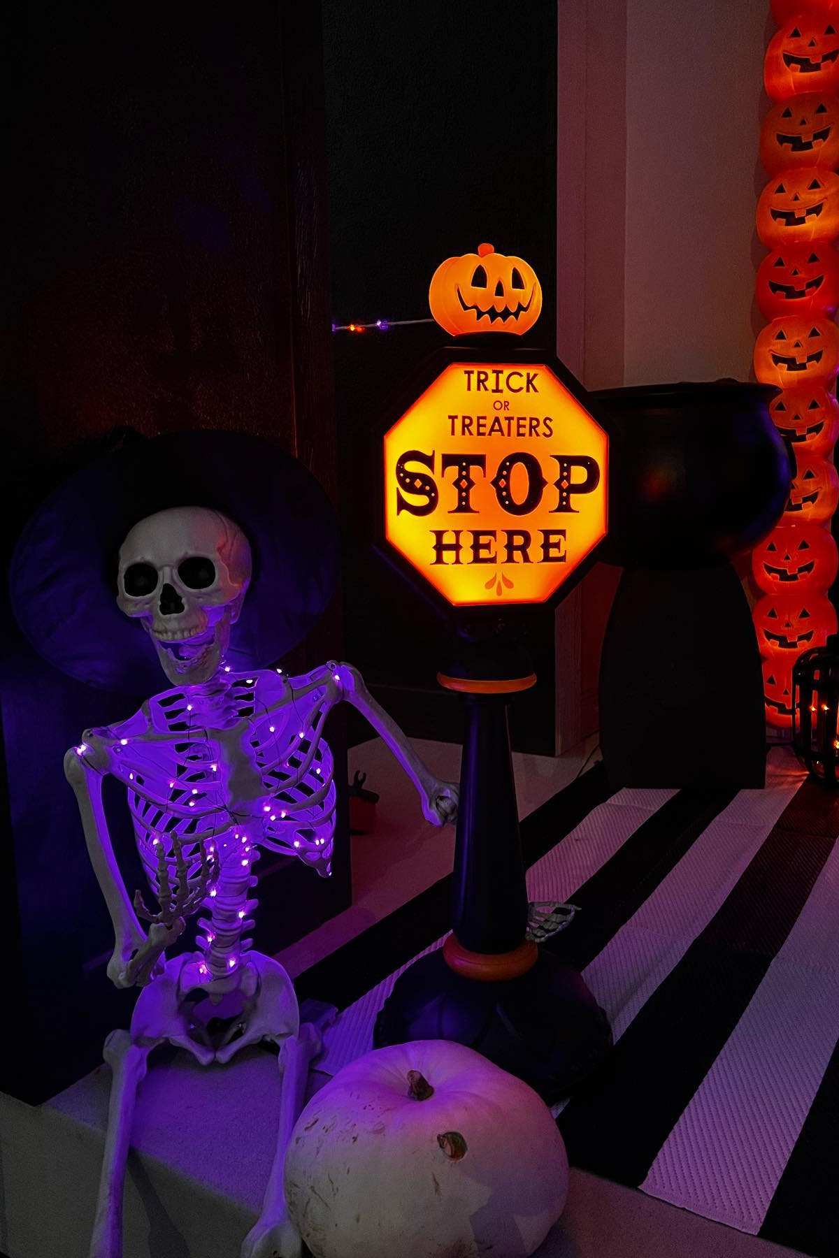 A plastic skeleton sits next to a glowing sign reading "Trick or Treaters Stop Here," with a pumpkin decoration on top and more pumpkin lights in the background.
