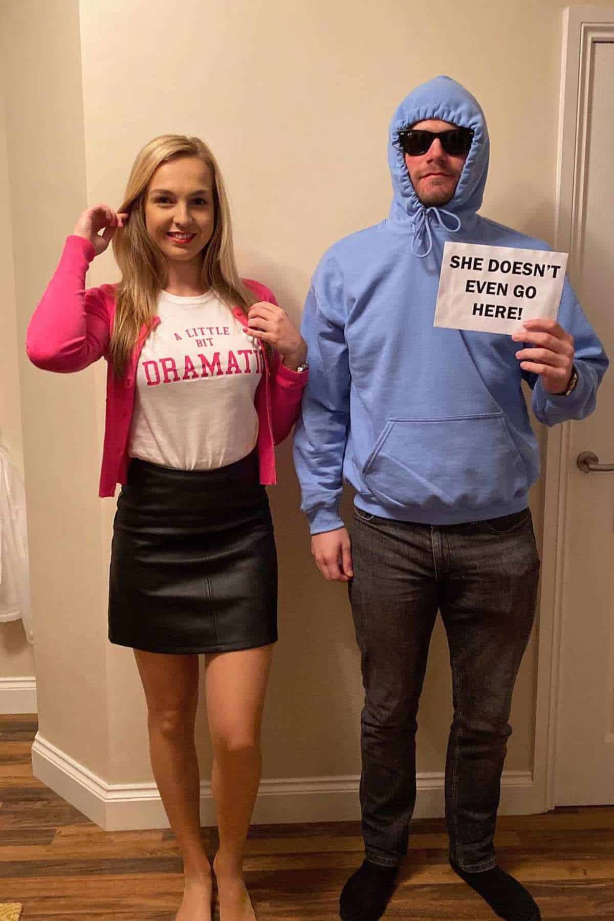 A woman in a pink cardigan and skirt stands next to a man in sunglasses and a blue hoodie holding a sign that says, "SHE DOESN'T EVEN GO HERE!.
