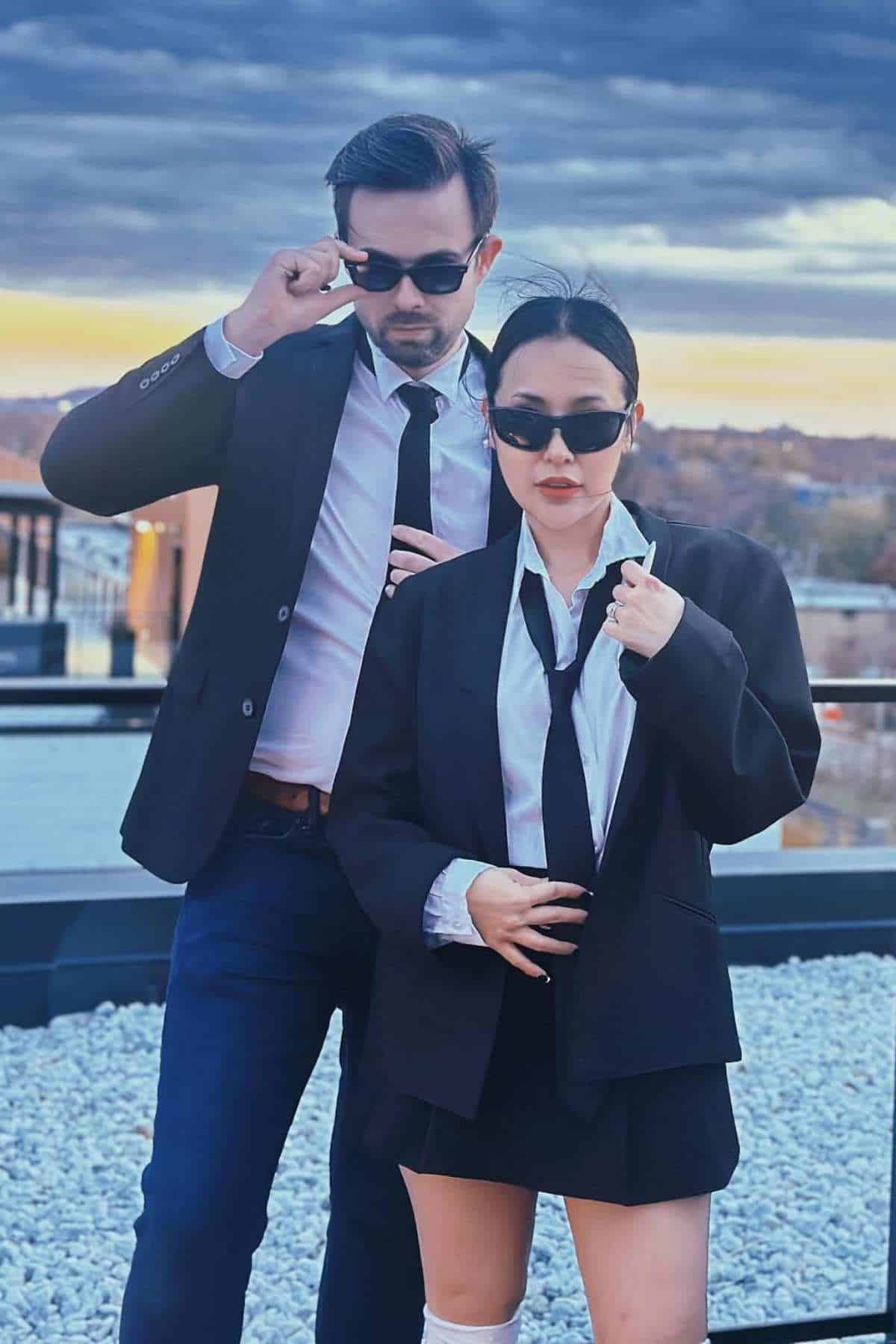 Two people in black suits, white shirts, and black sunglasses pose confidently on a rooftop with a cloudy sky and cityscape in the background.