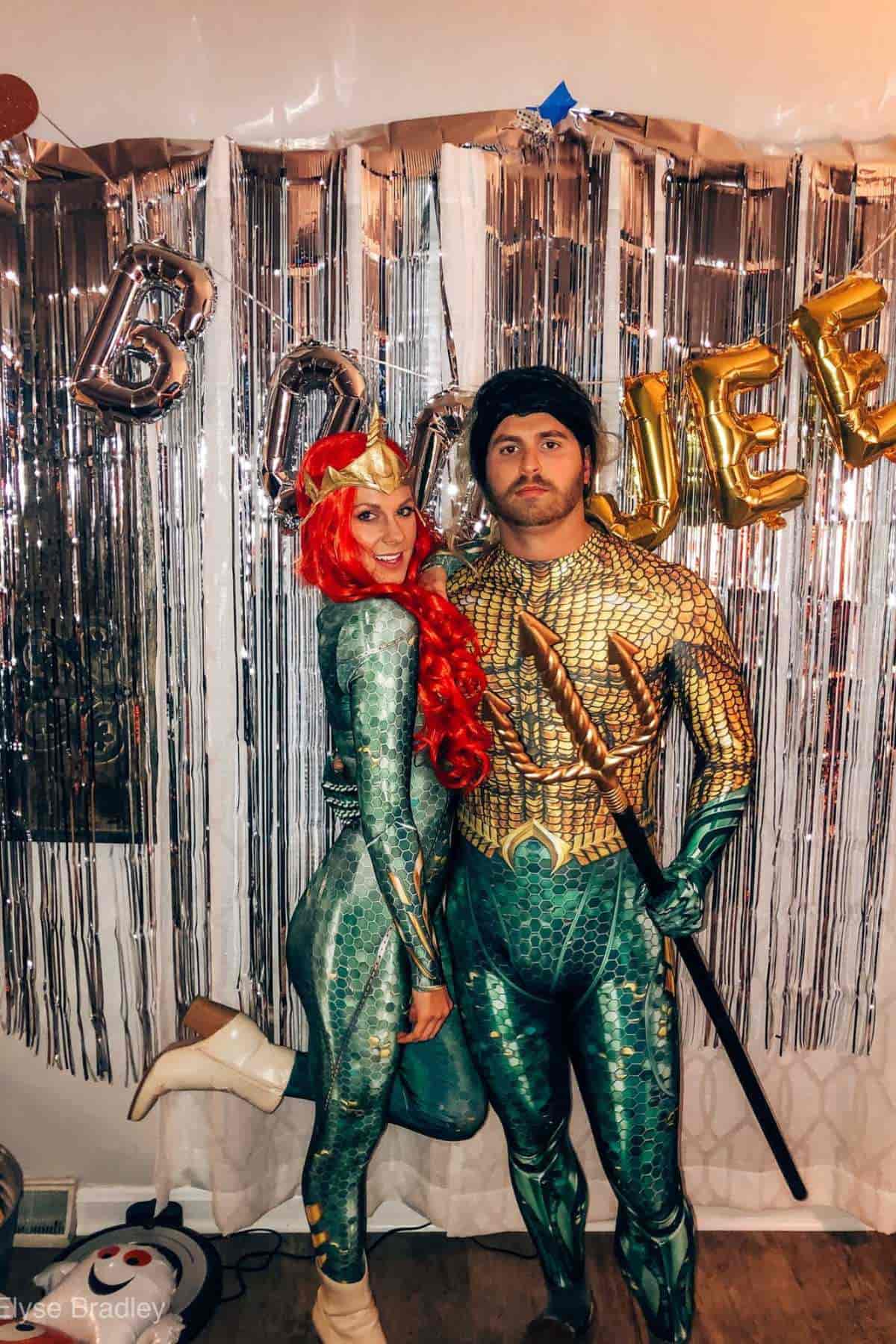 Two people in Aquaman-themed costumes pose in front of silver fringe streamers and balloon letters spelling "BOO" and "JEE." One person is holding a trident and the other has red hair.
