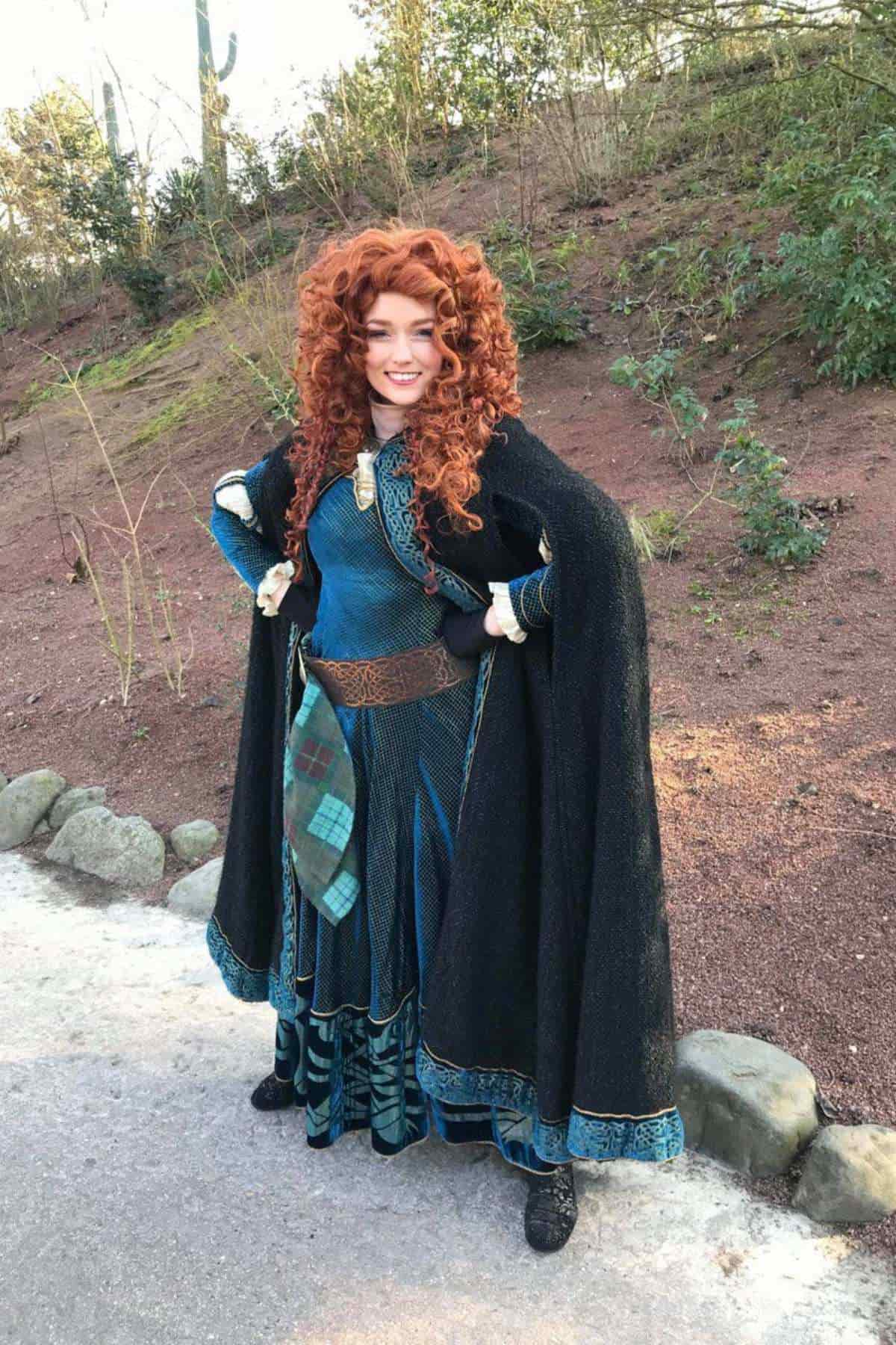 Person in a blue medieval-style dress with a tartan sash and a black cloak stands outdoors, hands on hips, smiling.