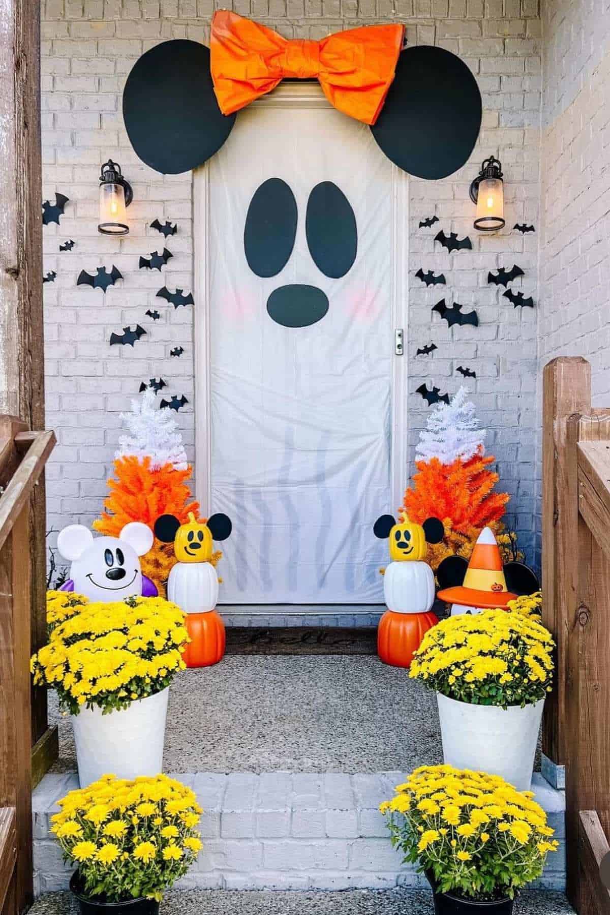 A front porch decorated with a Halloween Mickey Mouse theme, featuring a large bow, pumpkins, yellow flowers, bat cutouts, and white and orange trees.