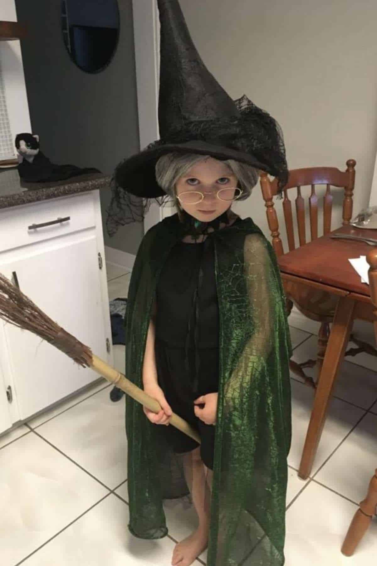 A child in a black dress, green cape, pointed witch hat, and glasses holds a broom, standing barefoot in a kitchen next to a wooden chair and table.