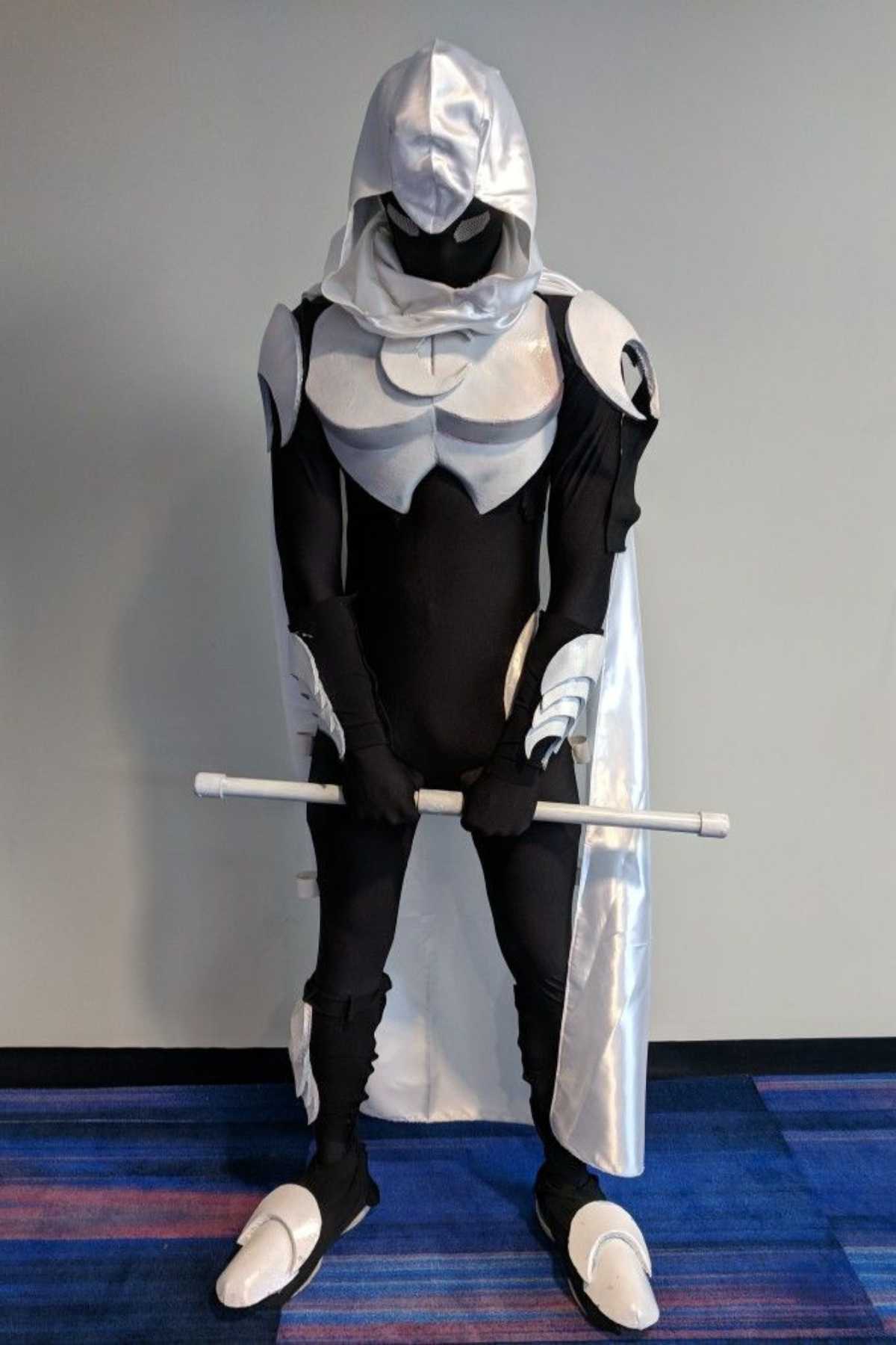 Person in a black and white superhero costume with armor, hood, cape, and staff, standing against a plain wall with blue and purple carpeted floor.