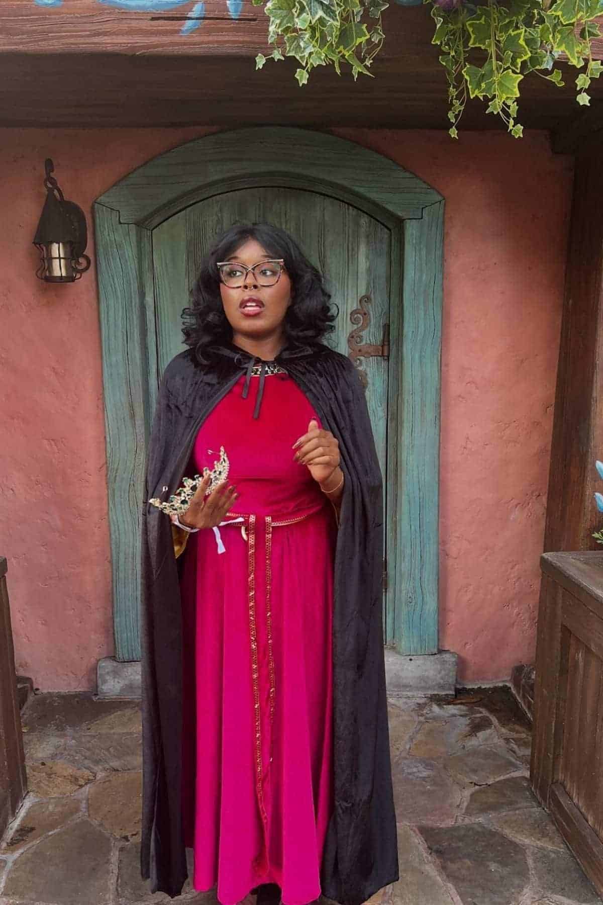 A person in a red dress and black cape stands in front of a rustic blue door, holding a gold crown and looking to the side.