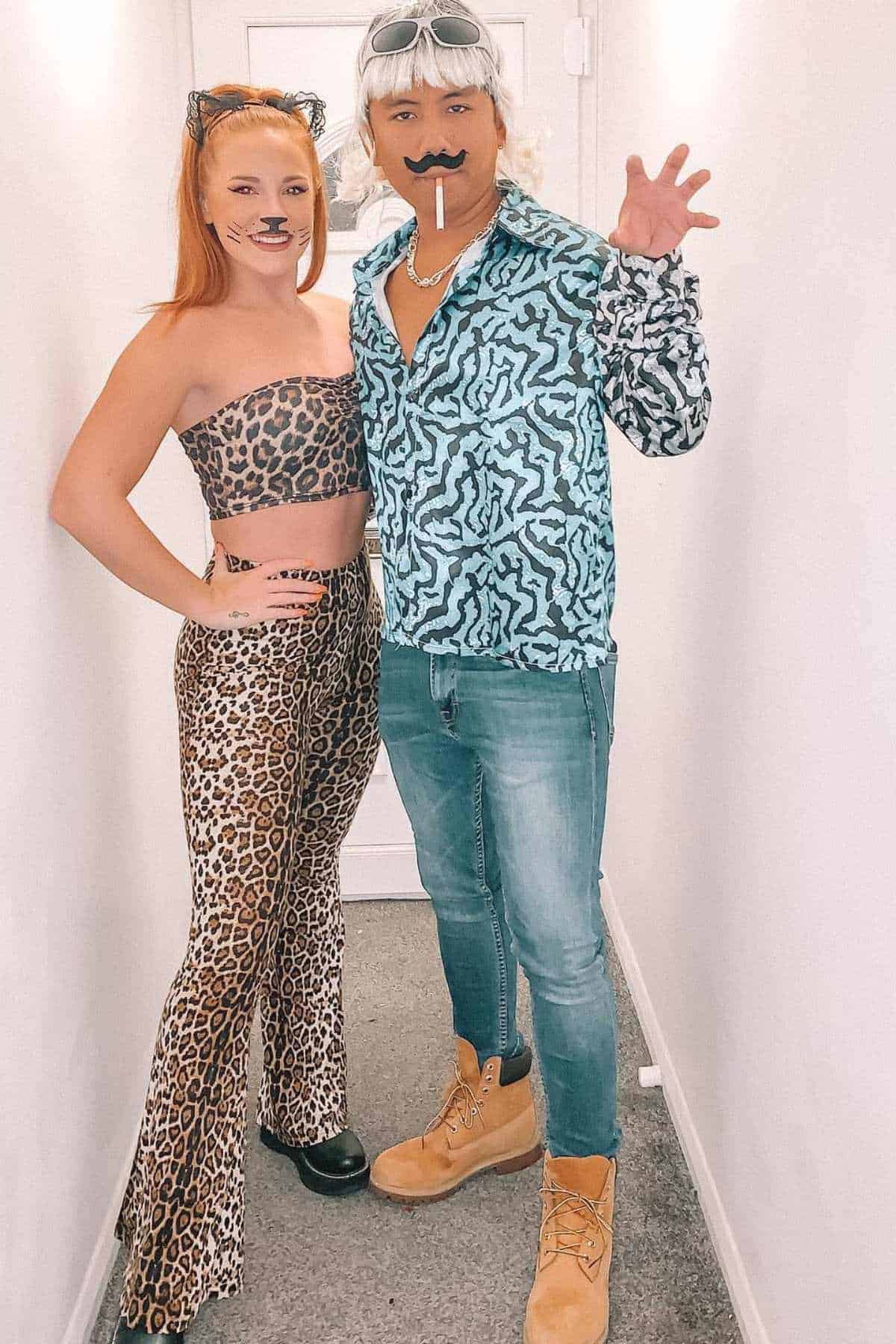 Two people pose in a hallway wearing costumes; one is dressed in leopard print with cat ears and face paint, the other in a patterned shirt, fake mustache, wig, and holding a fake cigarette.