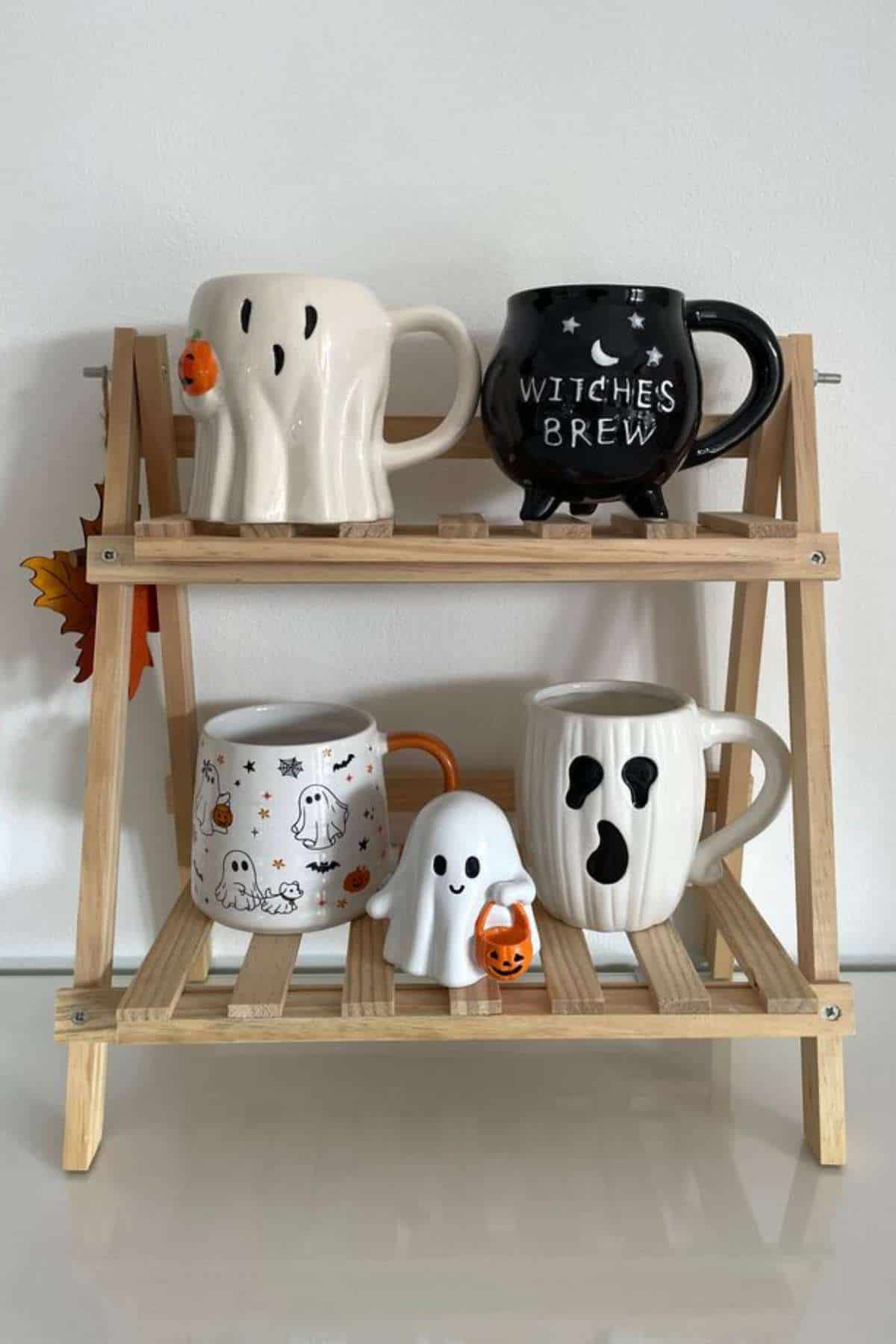 A wooden shelf displays five Halloween-themed mugs shaped like ghosts and a cauldron, with fall leaf decor on the side.