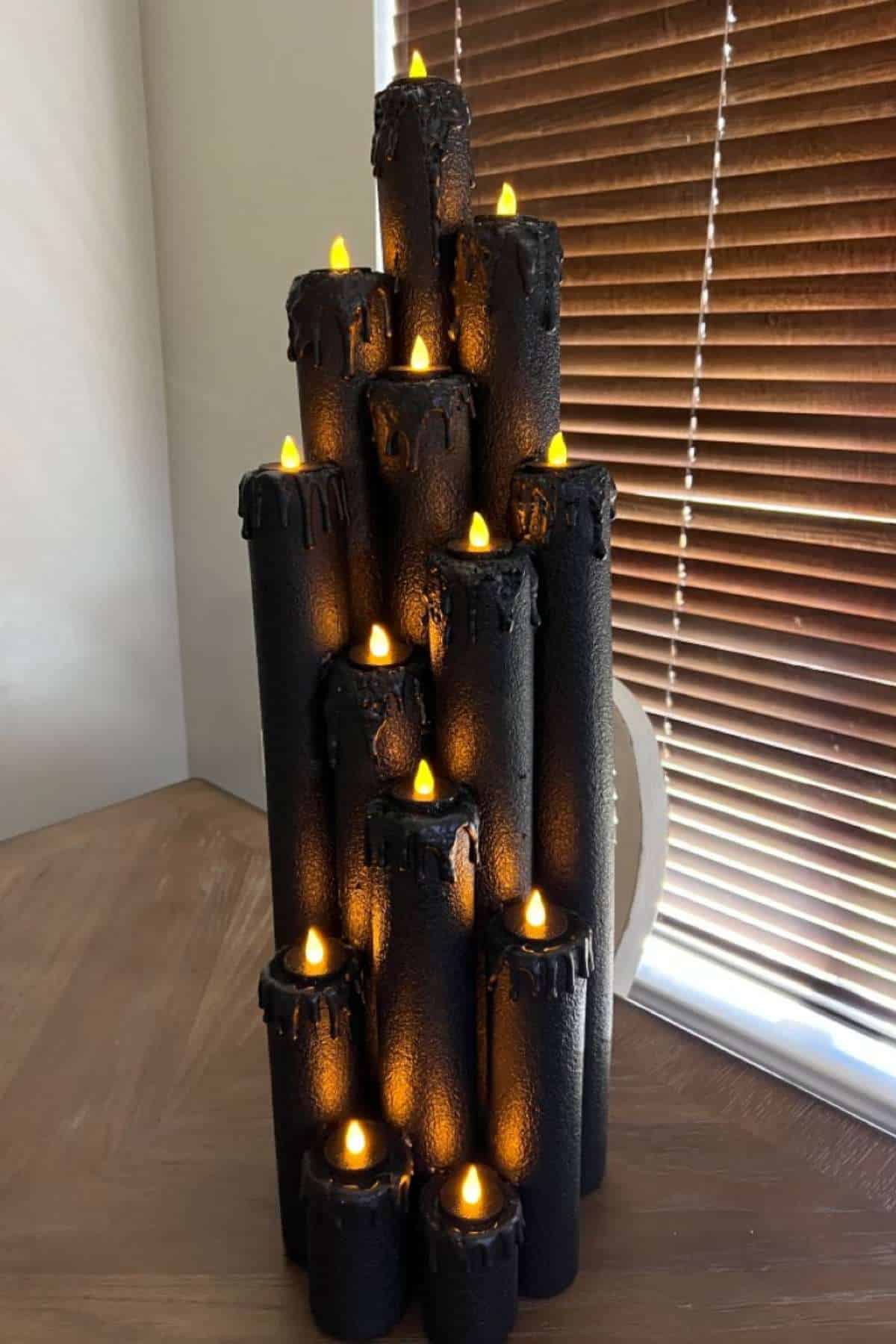 A group of black artificial candles with orange LED flames is arranged in a pyramid shape on a wooden table near window blinds.