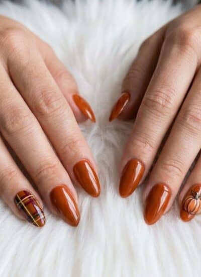 Two hands with long, almond-shaped nails painted brown; one nail on each hand features detailed plaid and spiderweb designs. Hands rest on a white fuzzy surface.