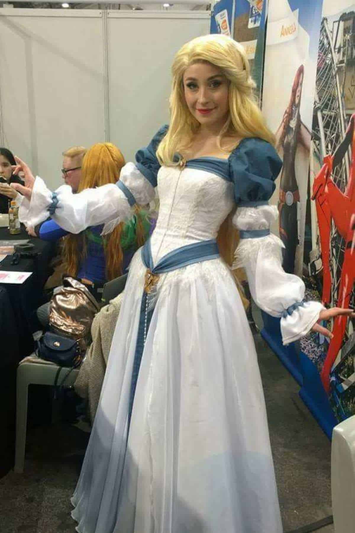 Person wearing a long, blue and white medieval-style dress with puffy sleeves and a blonde wig, posing at an indoor convention with booths and banners in the background.