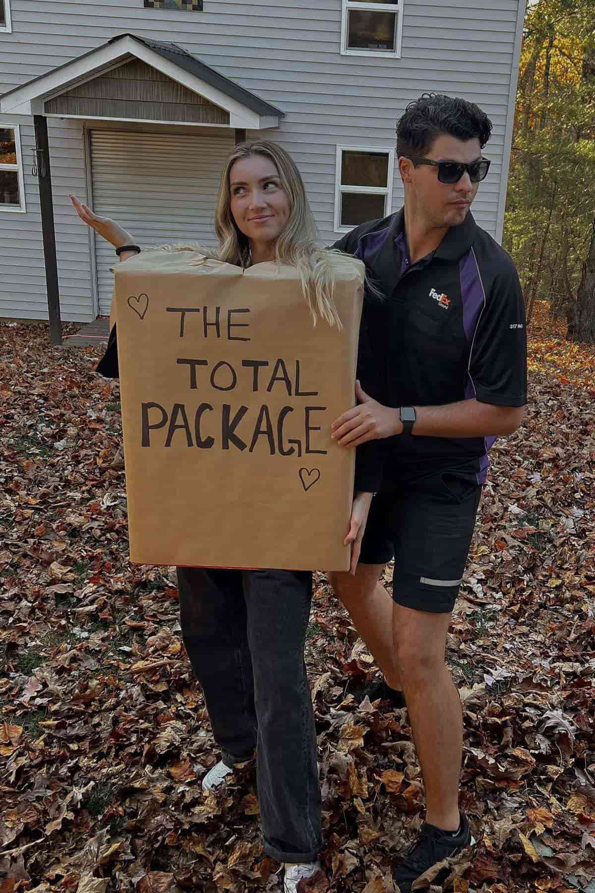 Two people stand outside; one is dressed as a FedEx delivery worker, and the other is wearing a large box labeled "THE TOTAL PACKAGE." They pose on fallen autumn leaves in front of a house.