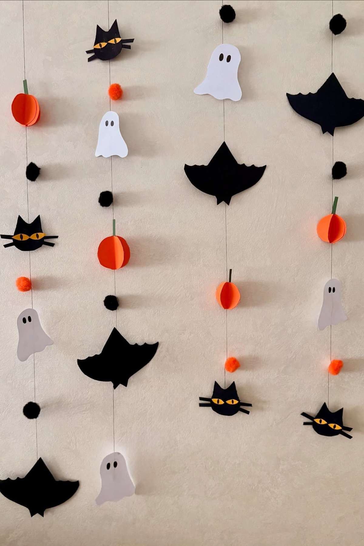 Paper decorations of bats, cats, ghosts, pumpkins, and pom-poms hang in vertical rows against a beige wall.