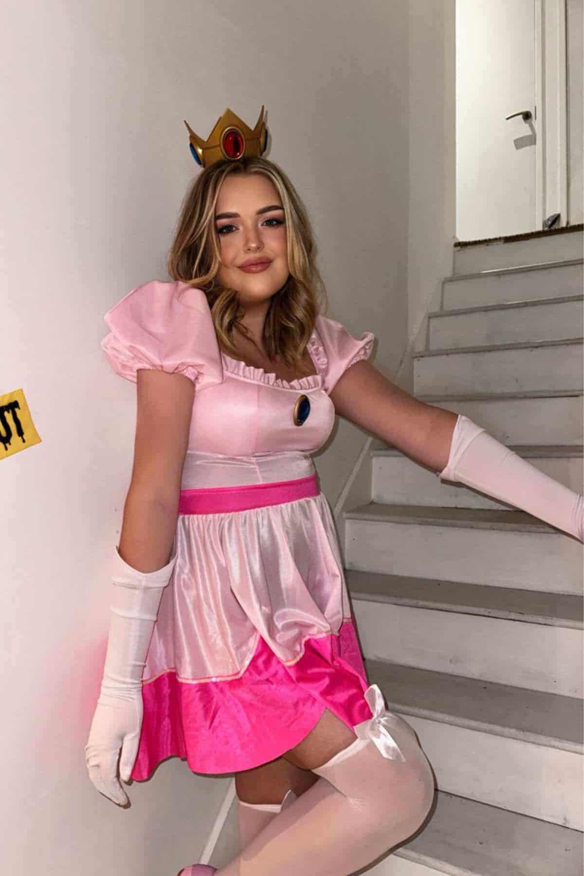 Person in a pink and white princess costume with a crown and gloves poses on a white staircase.
