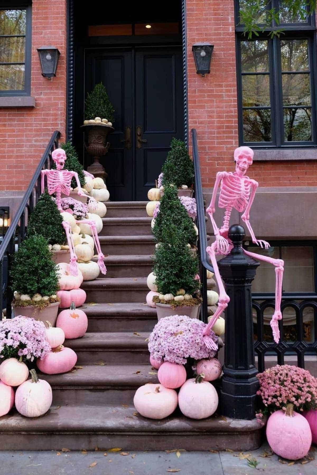 Two pink skeleton decorations sit among pink and white pumpkins and potted plants on the steps and railing of a brick townhouse.