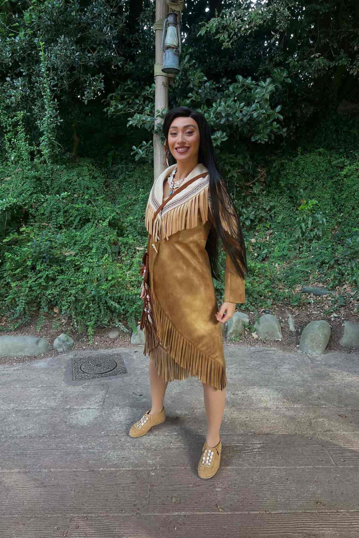 Person wearing a fringed brown dress and moccasins stands outdoors on a path with greenery in the background, smiling at the camera.