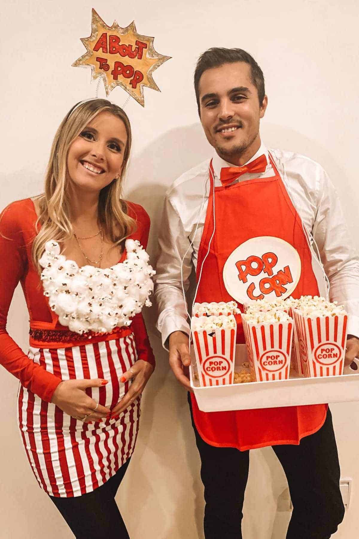 Two people in coordinated popcorn-themed costumes; one wears a "About to Pop" headband and popcorn-decorated shirt, the other wears a "Popcorn" apron and holds a tray of popcorn containers.