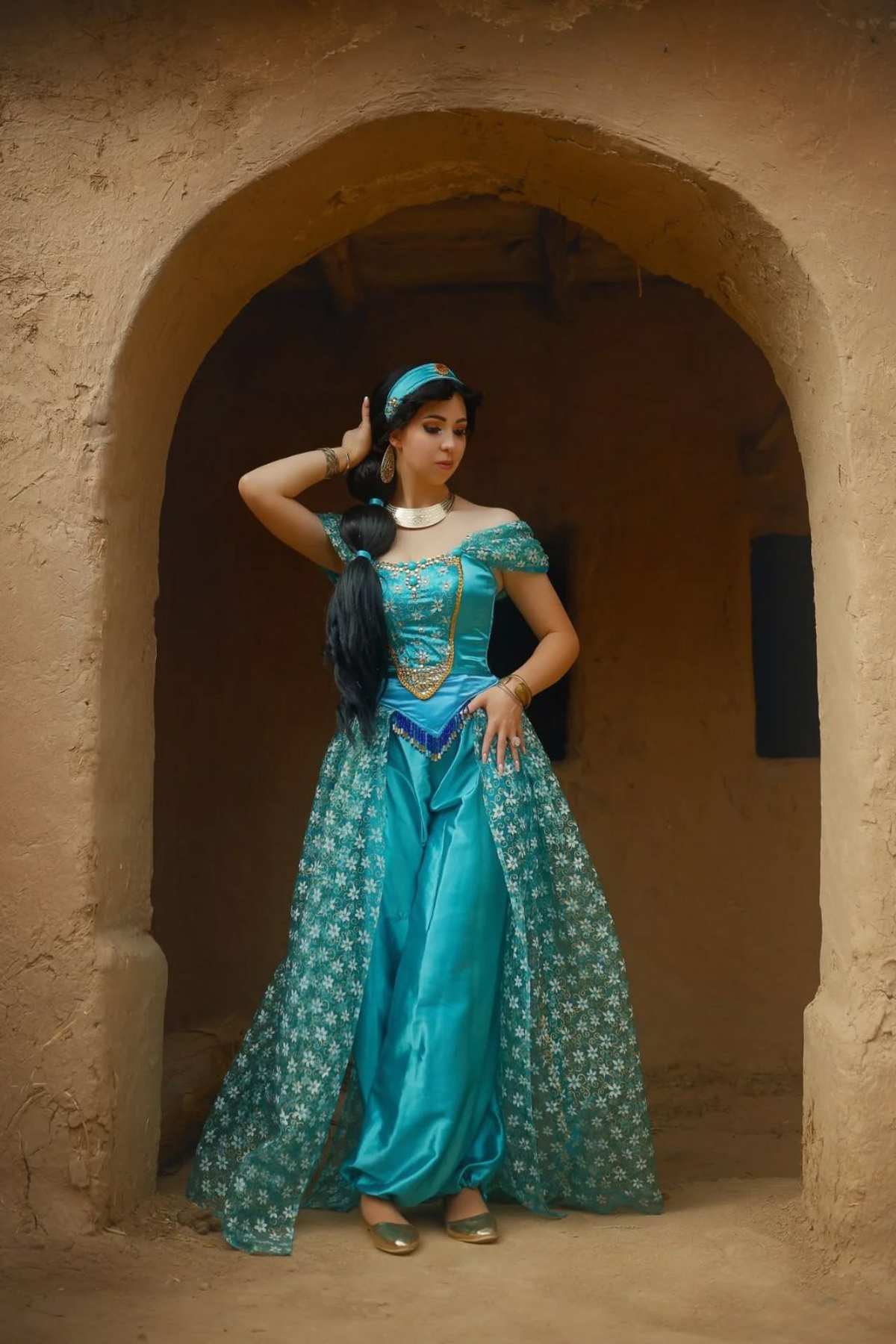Woman in a turquoise, off-shoulder, princess-style costume poses under a rustic archway with her hand on her head and hip.