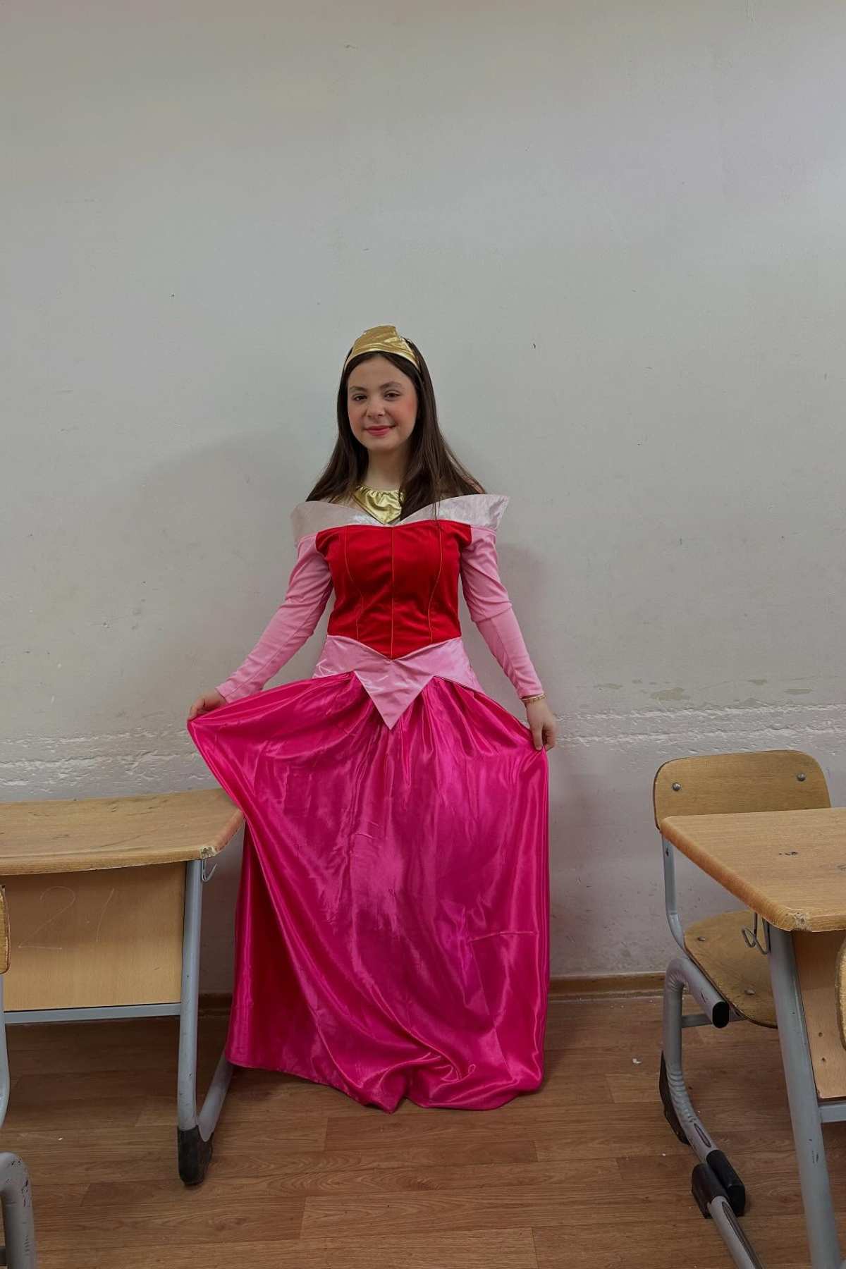 A person in a pink princess costume stands between two wooden desks in a classroom with a plain white wall background.