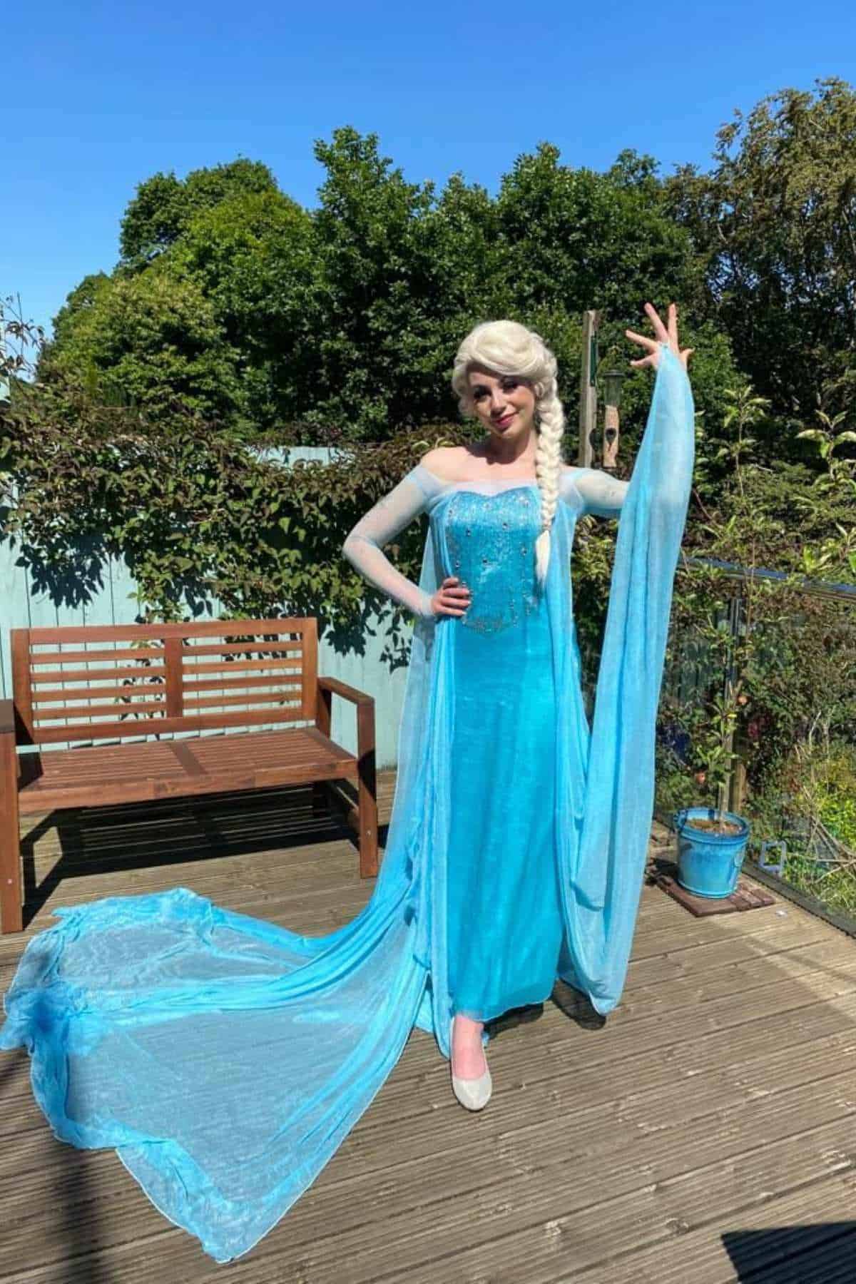 Person in an Elsa costume from Frozen stands on a wooden deck, posing with one arm raised, with greenery and a bench in the background.