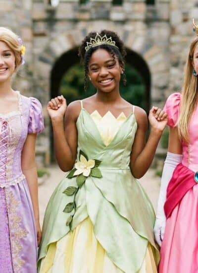 Three women in colorful princess dresses and crowns stand smiling in front of a stone castle and garden.