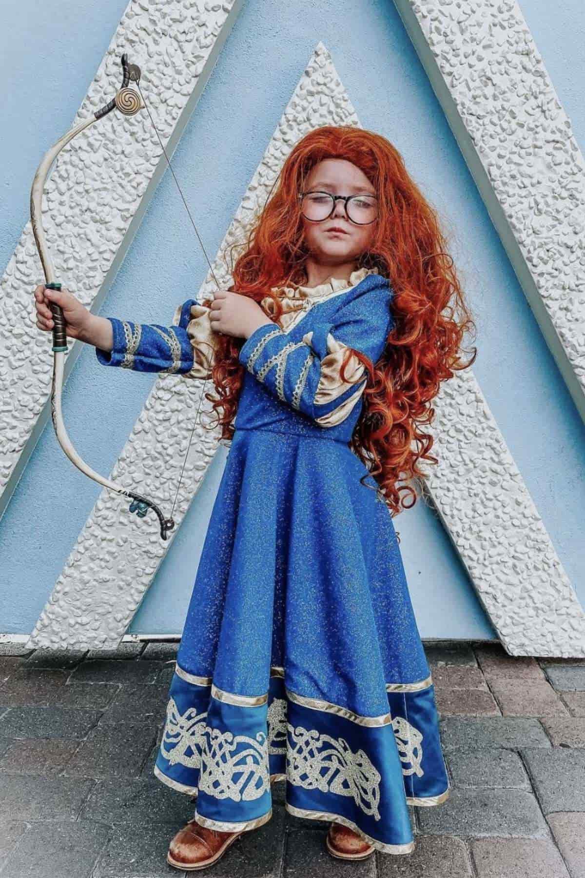 Child in a blue and gold dress with long curly red hair and glasses holds a bow, standing in front of a textured wall with a geometric pattern.
