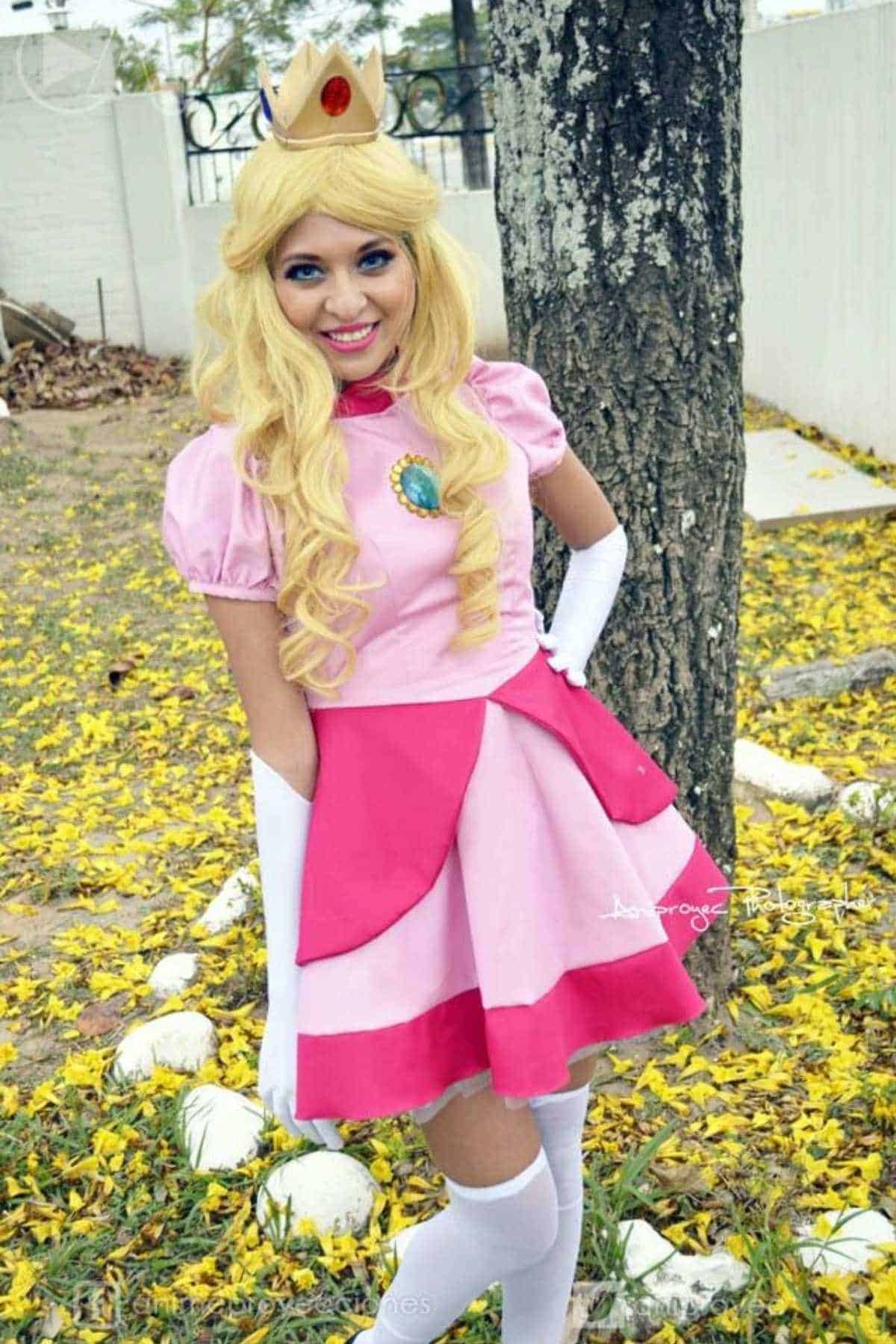 Person wearing a pink and peach princess costume with a crown, long blond wig, and white gloves, standing outdoors near a tree and yellow scattered petals.