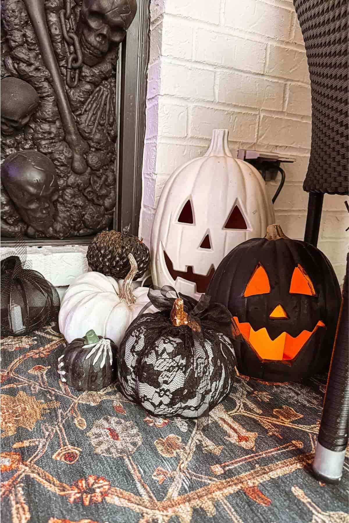 Several decorative pumpkins, including black, white, and lace-covered ones, are arranged on a patterned rug beside a fireplace with a skeleton relief. Two pumpkins have carved faces and are lit inside.