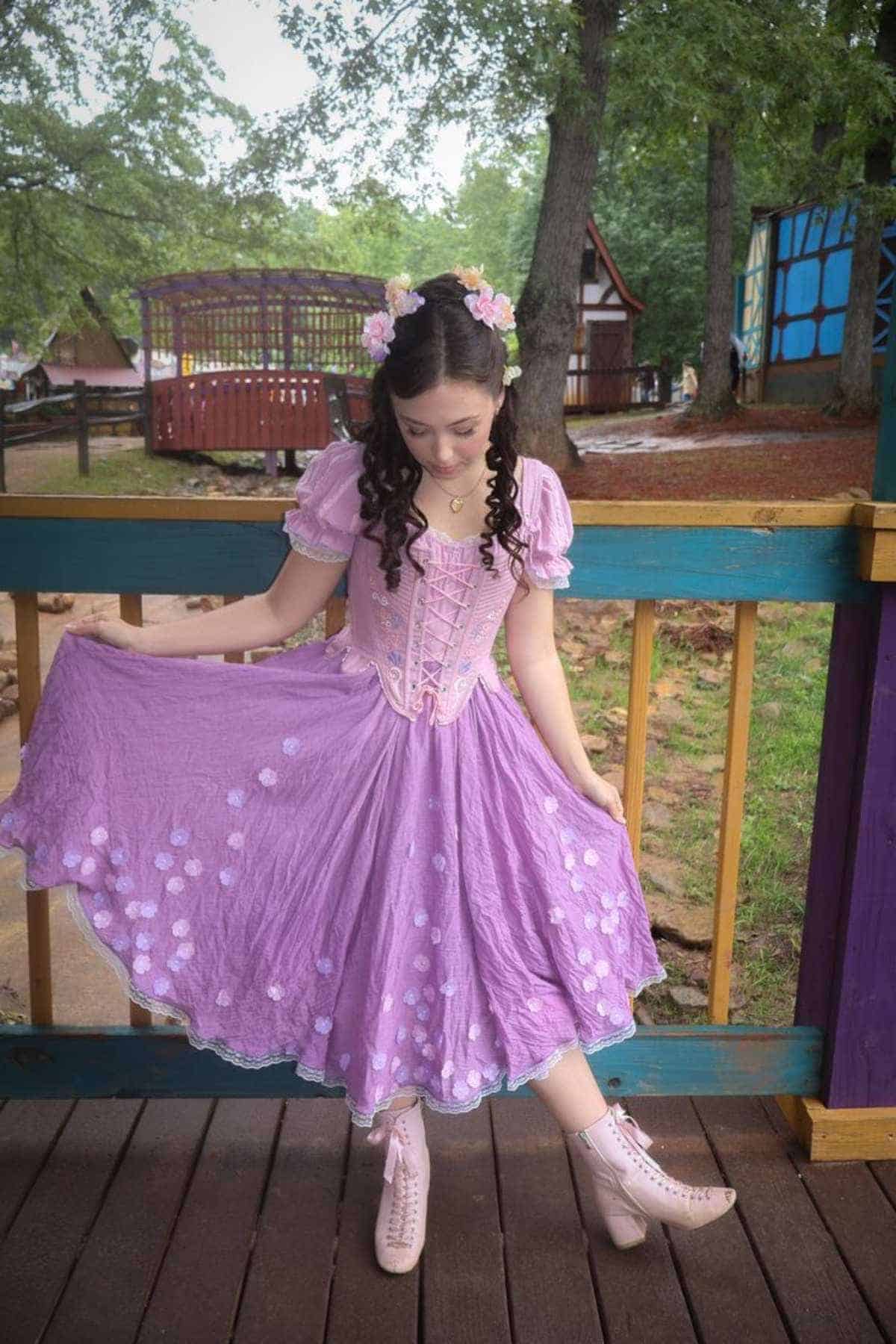 Young woman in a purple dress with floral details stands on a wooden deck, holding out her skirt. She wears boots and a flower crown, with trees and wooden structures in the background.