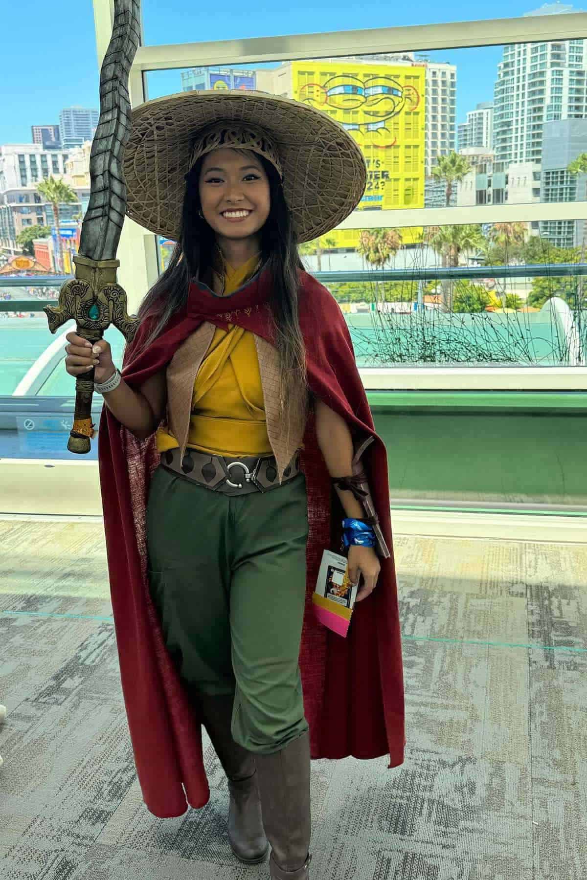 Person in cosplay outfit with a wide-brimmed hat, red cape, green pants, brown boots, and holding a large sword, standing indoors at a convention with city buildings in the background.