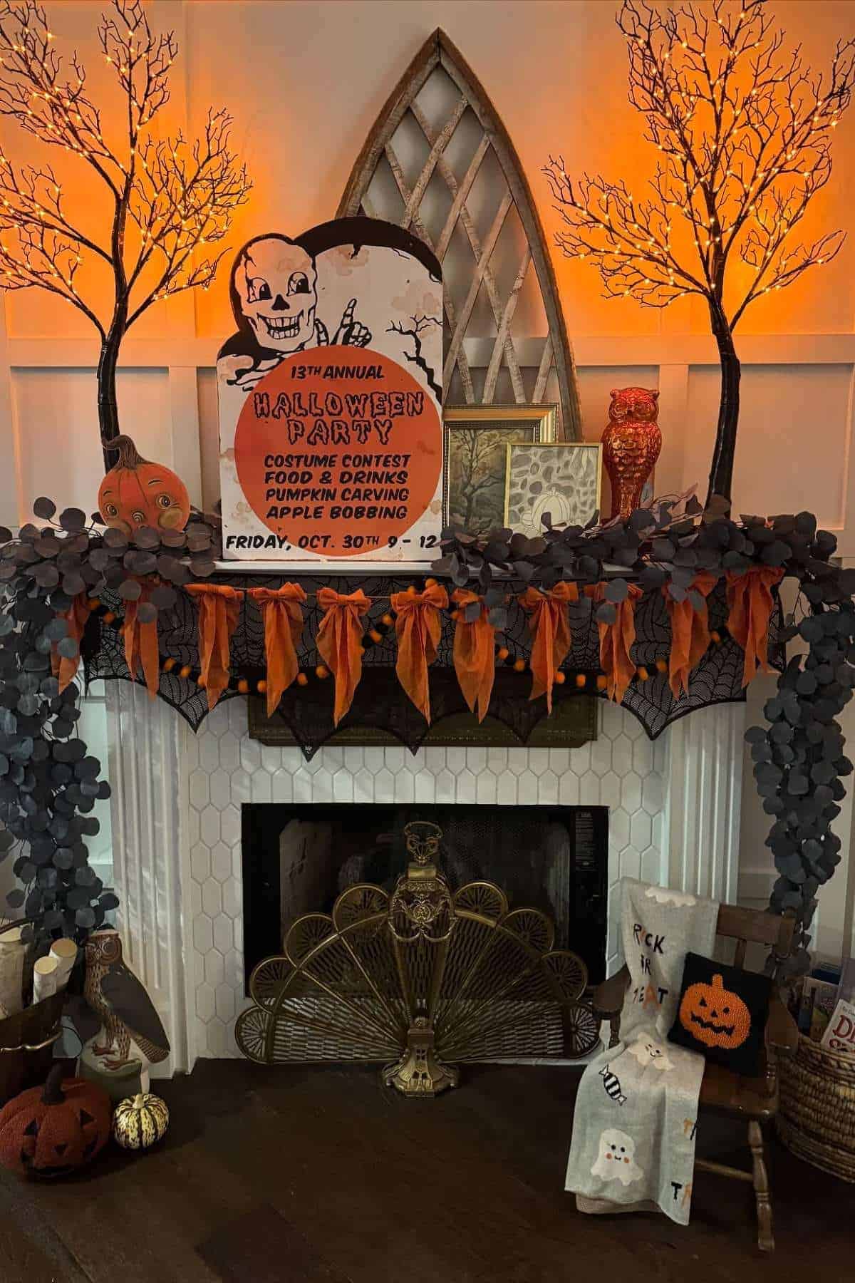 A Halloween-themed mantel with orange lights, decorative trees, a contest sign, pumpkins, and festive garlands above a white fireplace. Pillows and a pumpkin bucket sit on a chair nearby.