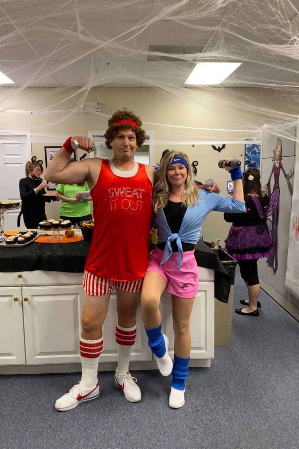 Two people in 1980s-style workout costumes pose and flex their arms at a party, with food on the counter and spider web decorations in the background.