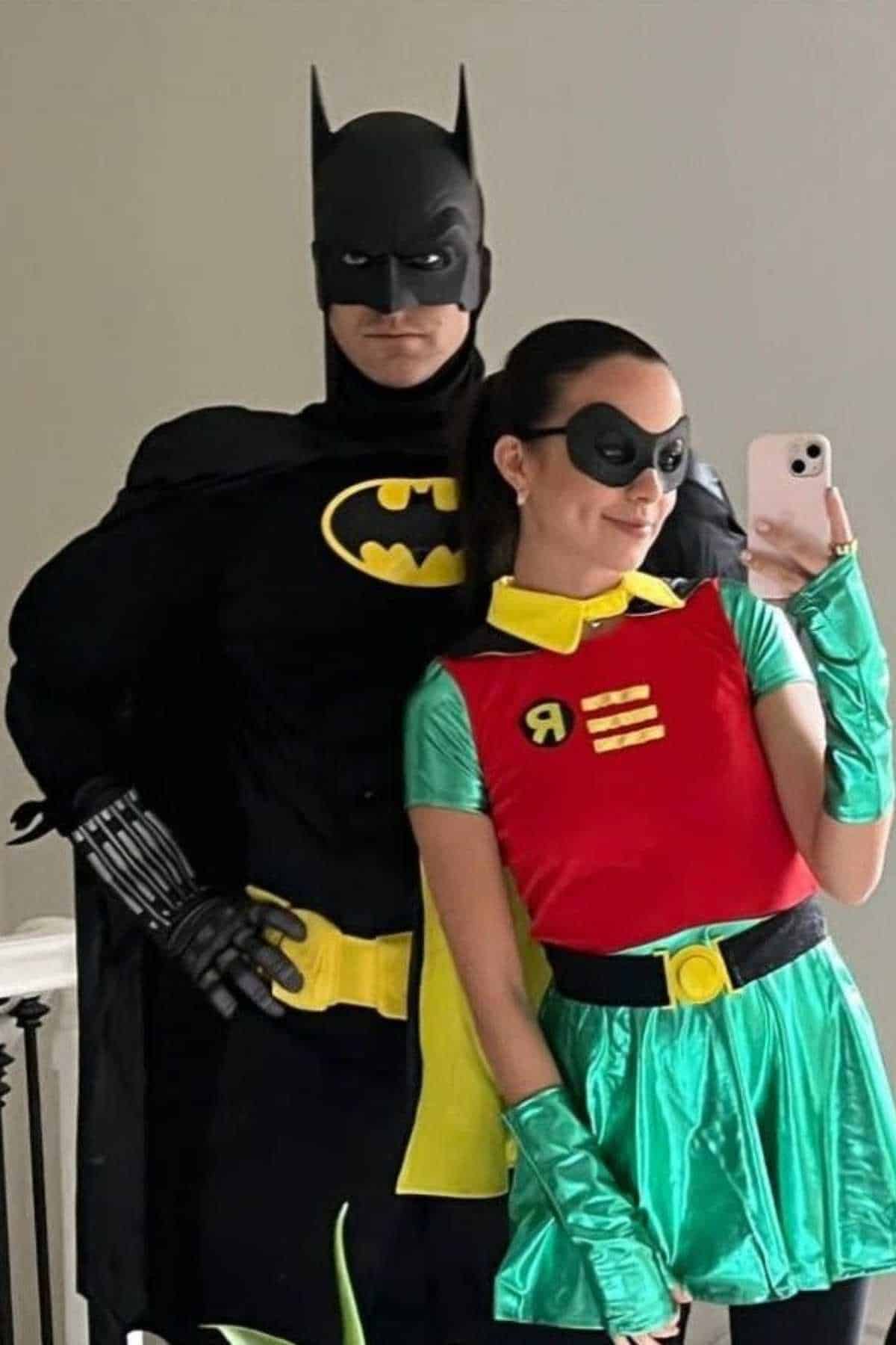 Two people wearing Batman and Robin costumes stand side by side and pose for a selfie in front of a mirror.