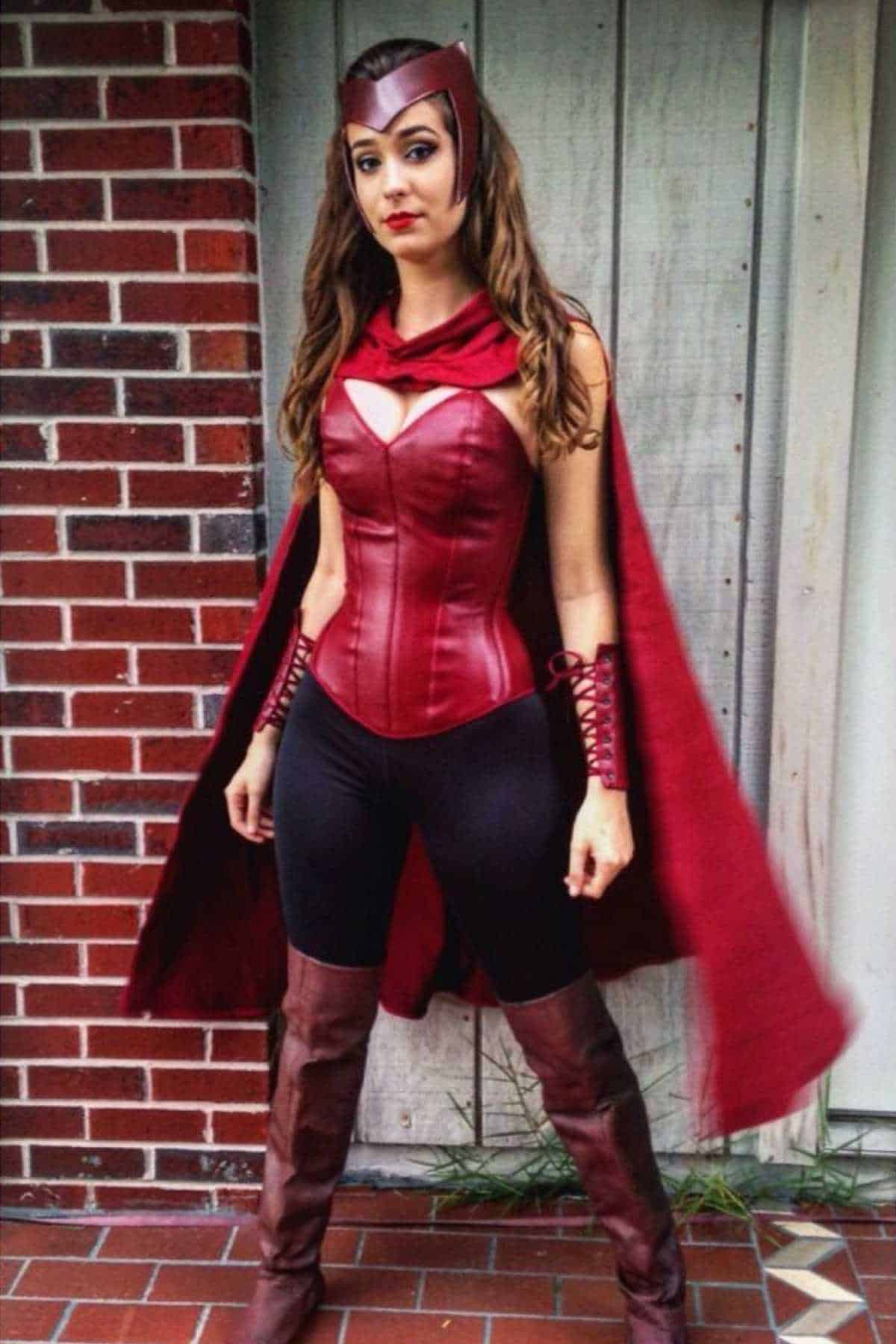 Person in a red and maroon superhero costume with a cape, headpiece, corset, bracers, and boots, standing in front of a brick and wooden wall.