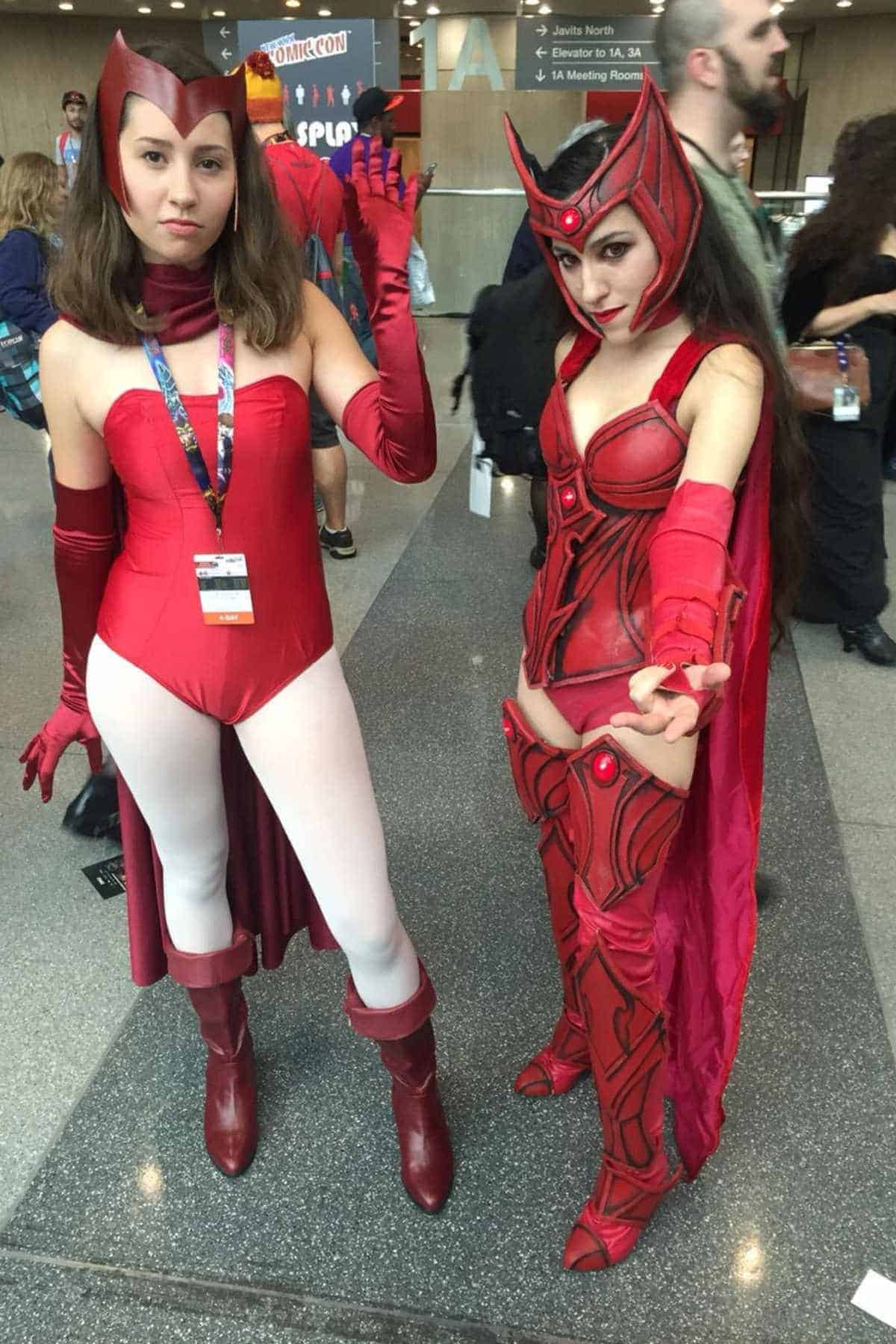 Two people at a convention dressed as the Scarlet Witch from Marvel Comics, wearing red costumes with headdresses and gloves, standing on a polished floor with other attendees in the background.