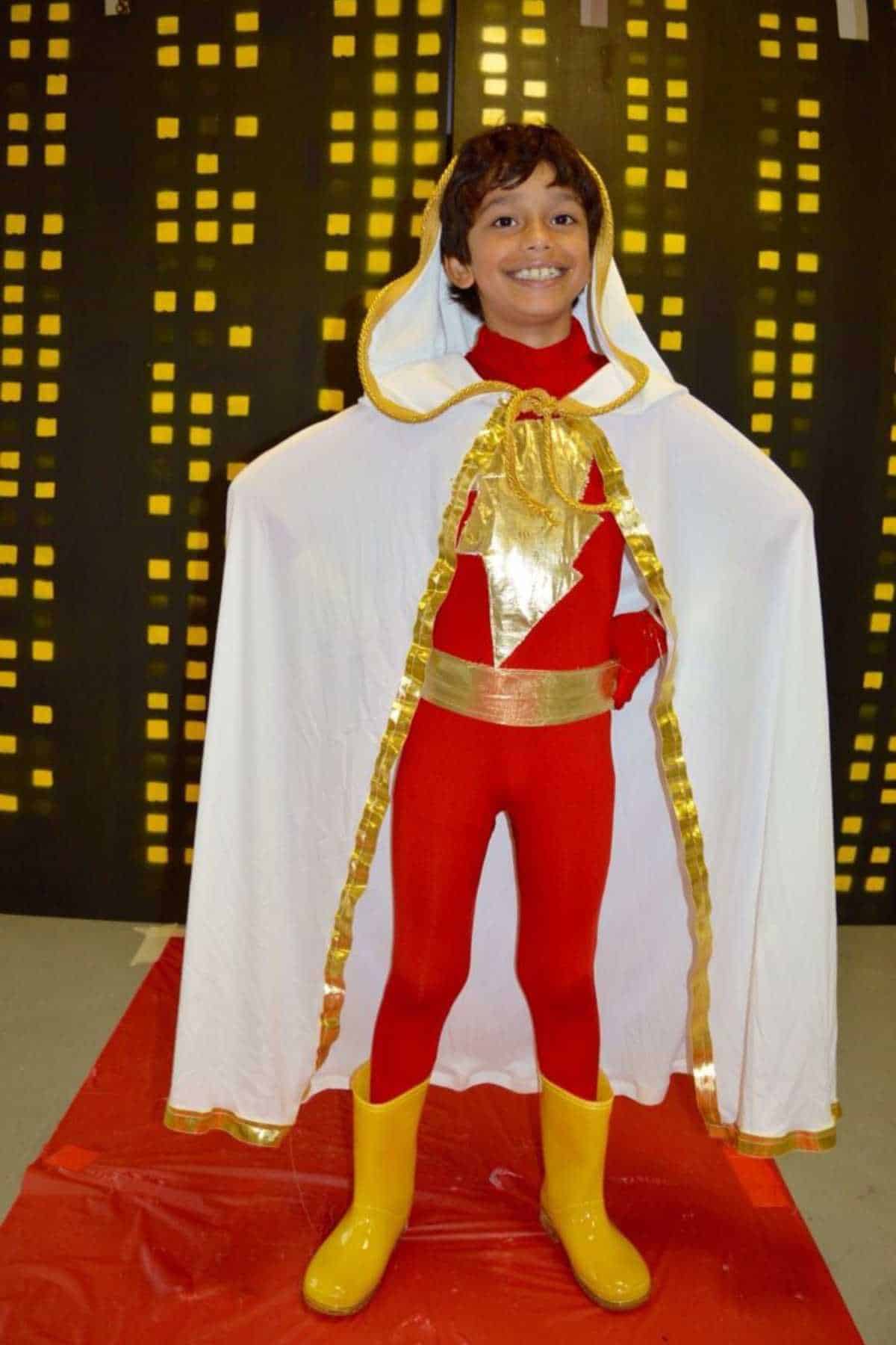 Child in a red and yellow superhero costume with a white cape stands smiling in front of a backdrop resembling city buildings.