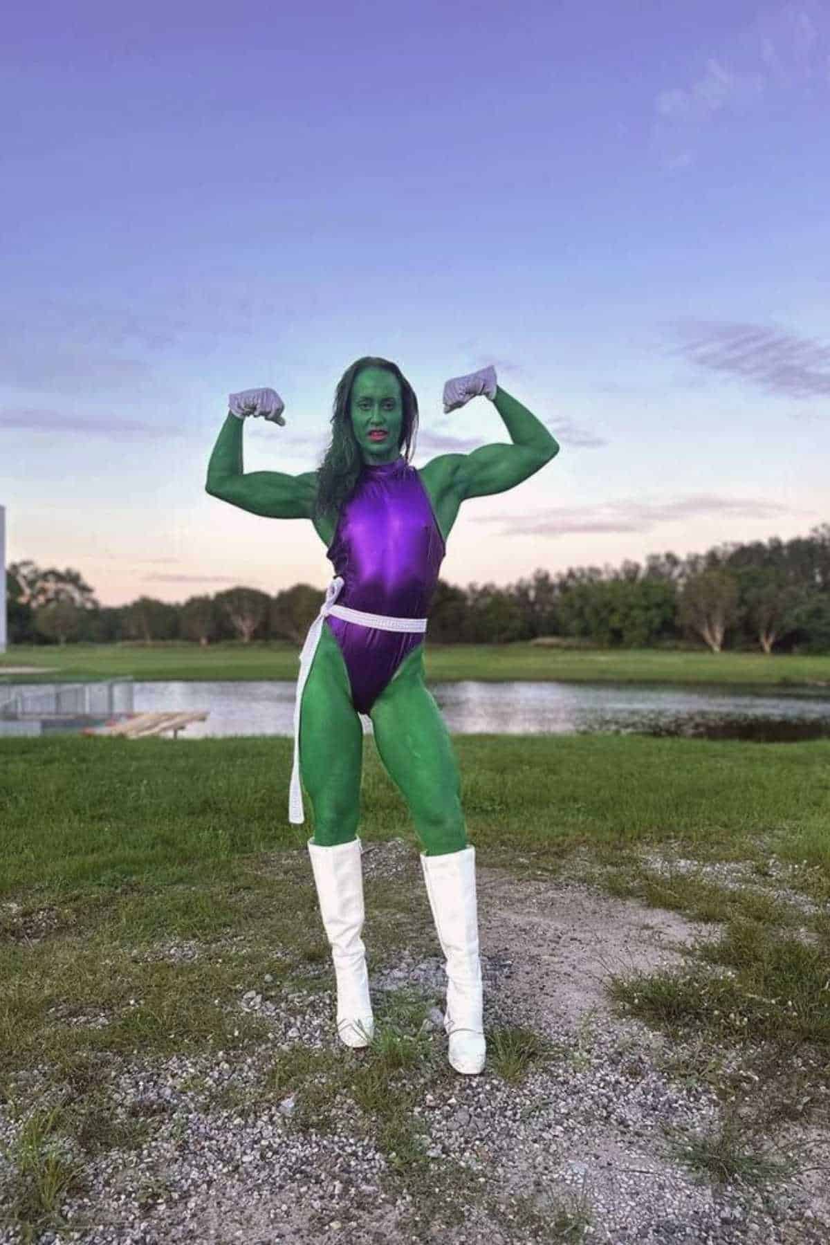 Person dressed as She-Hulk stands outdoors on grass, flexing muscles, wearing a purple costume, white boots, and gloves, with green body paint and a pond in the background.