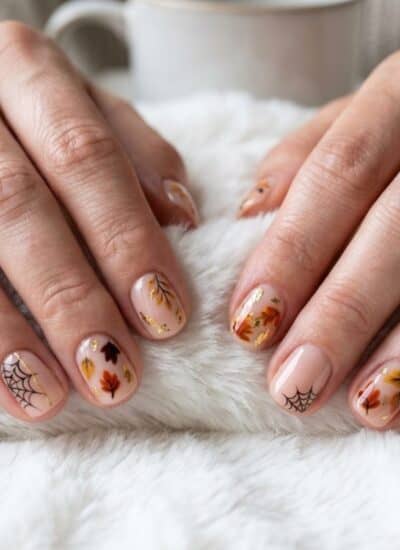 Close-up of hands with autumn-themed nail art, featuring leaves and spiderweb designs, resting on a white fuzzy blanket; a white cup is in the background.