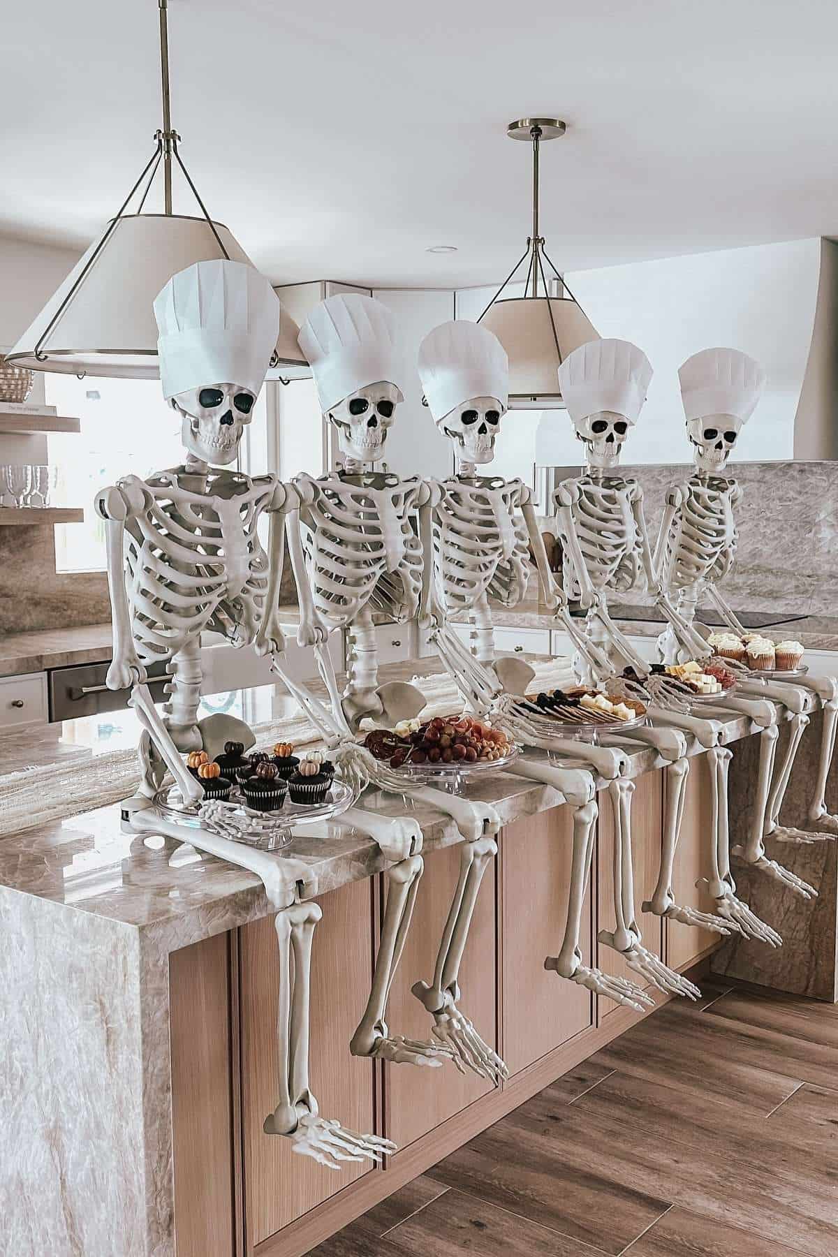 Five skeletons wearing chef hats sit on a kitchen counter with plates of desserts and fruit in front of them.