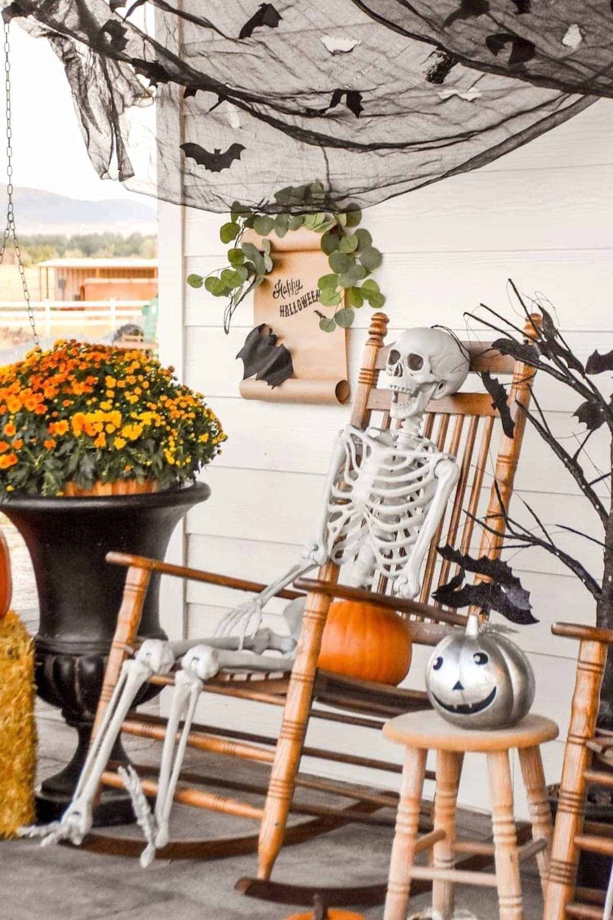 A skeleton sits in a wooden rocking chair on a porch decorated with flowers, pumpkins, black netting, and a "Happy Halloween" sign.